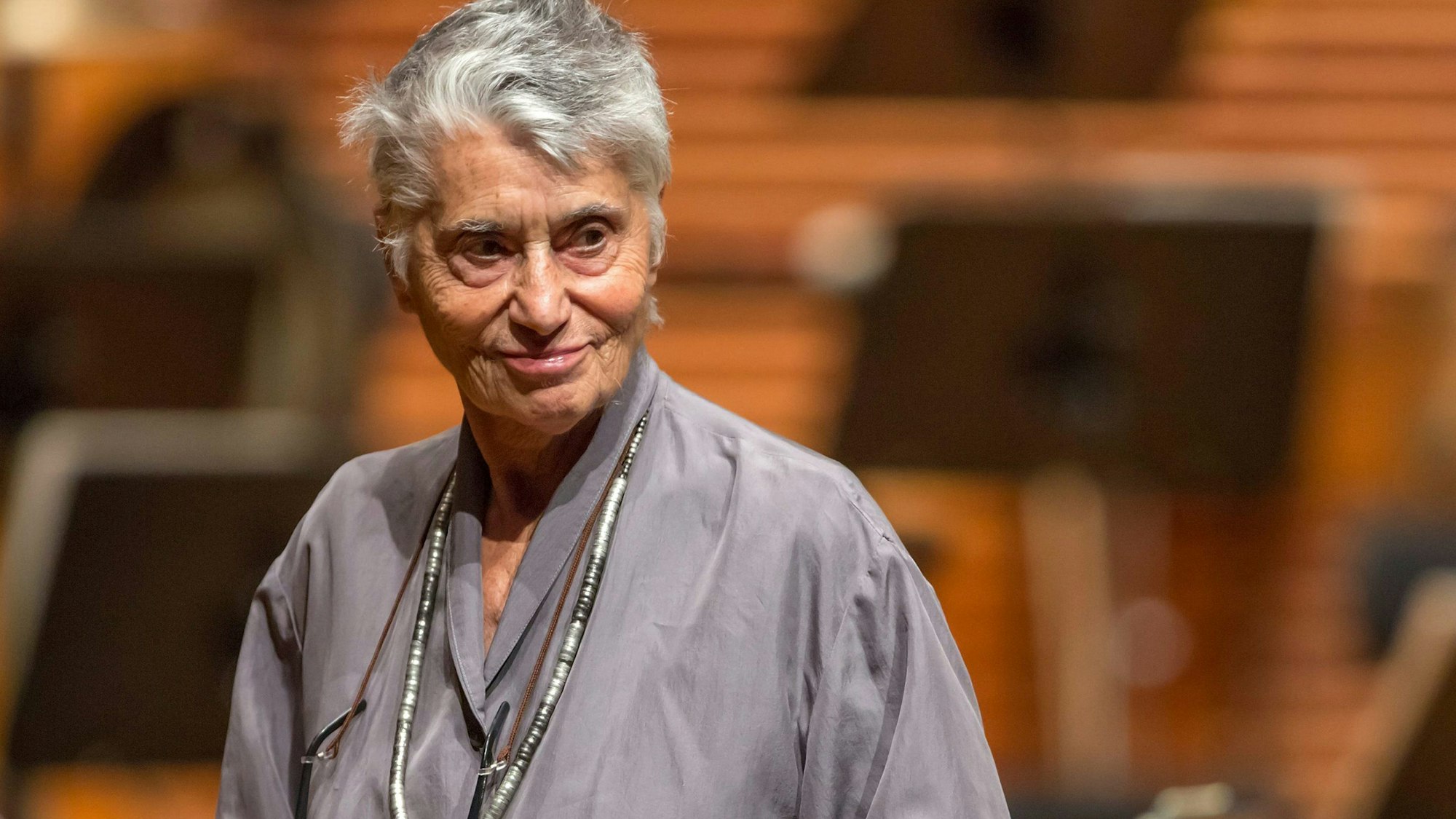 Ruth Klüger hat weiße Haare und ist betagt, sie trägt ein graues Hemd und eine schwarz-weiß-graue Kette.