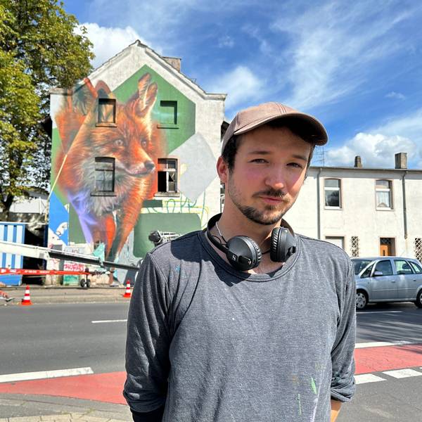 Ein junger Mann mit Käppi und Kopfhörern um den Hals steht an einer Straße. Im Hintergrund ist ein Gebäude zu sehen, auf dem ein großer Fuchs prangt.