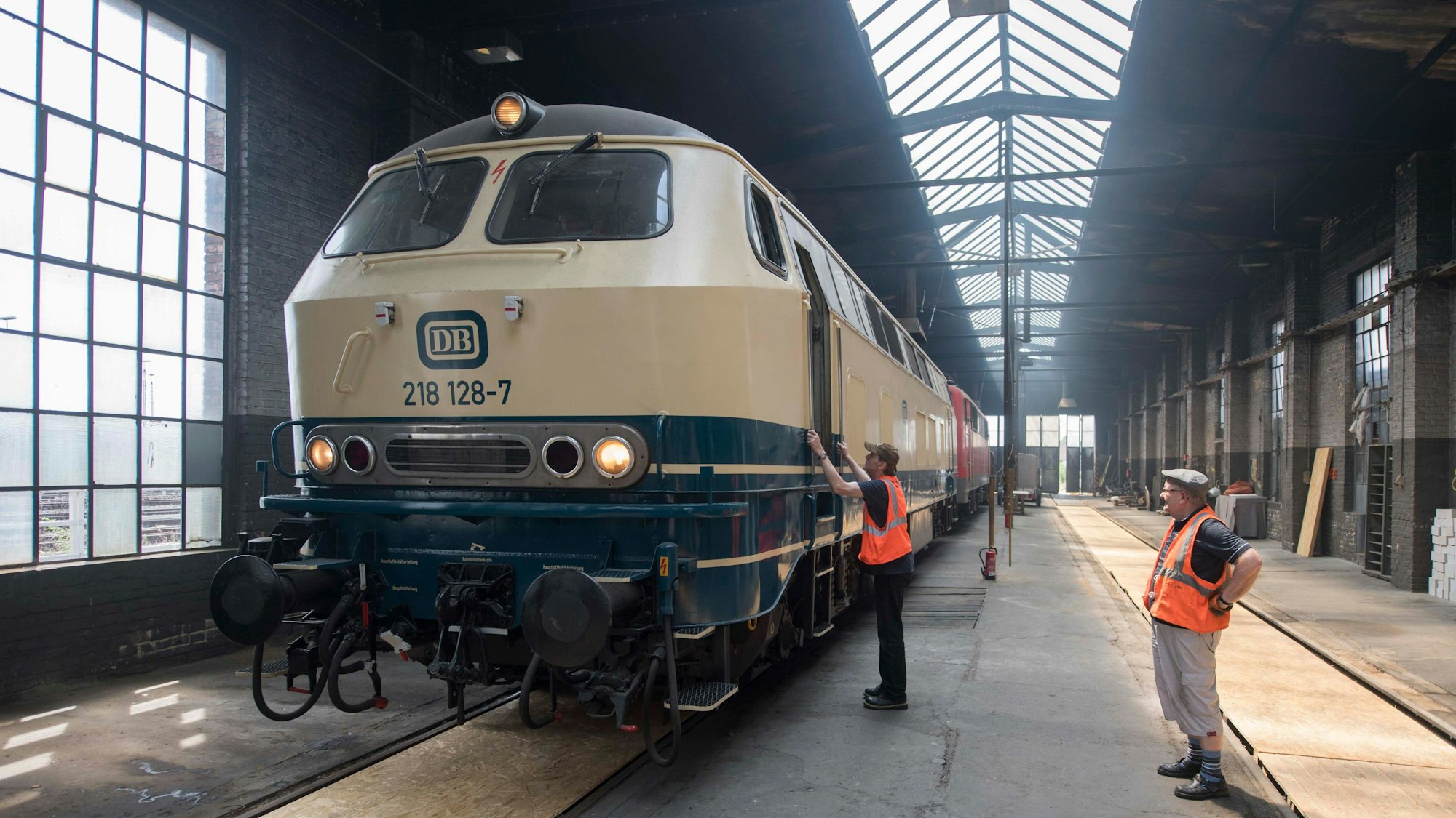 Zwei Männer in orangen Warnwesten stehen in einer großen Halle. Der Linke steht an einer historischen Lokomotive.