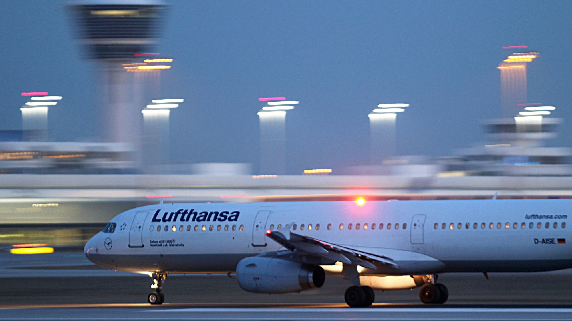 Ein Airbus A321 der deutschen Fluggesellschaft Lufthansa startet auf einem Flughafen. Im Hintergrund sind verschwommen Scheinwerfer der Startbahn und der Flughafentower zu sehen. (Symbolbild)