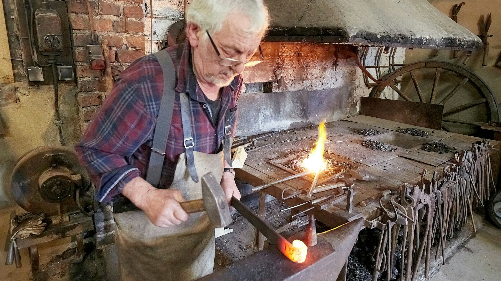 Der Schmied Stefan Pütz steht an einem Amboss. In der linken Hand hält er ein glühendes Eisen, in der rechten einen Hammer. Er trägt ein kariertes Hemd und eine Lederschürze. Im Hintergrund sind Werkzeuge und eine Feuerstelle zu sehen.