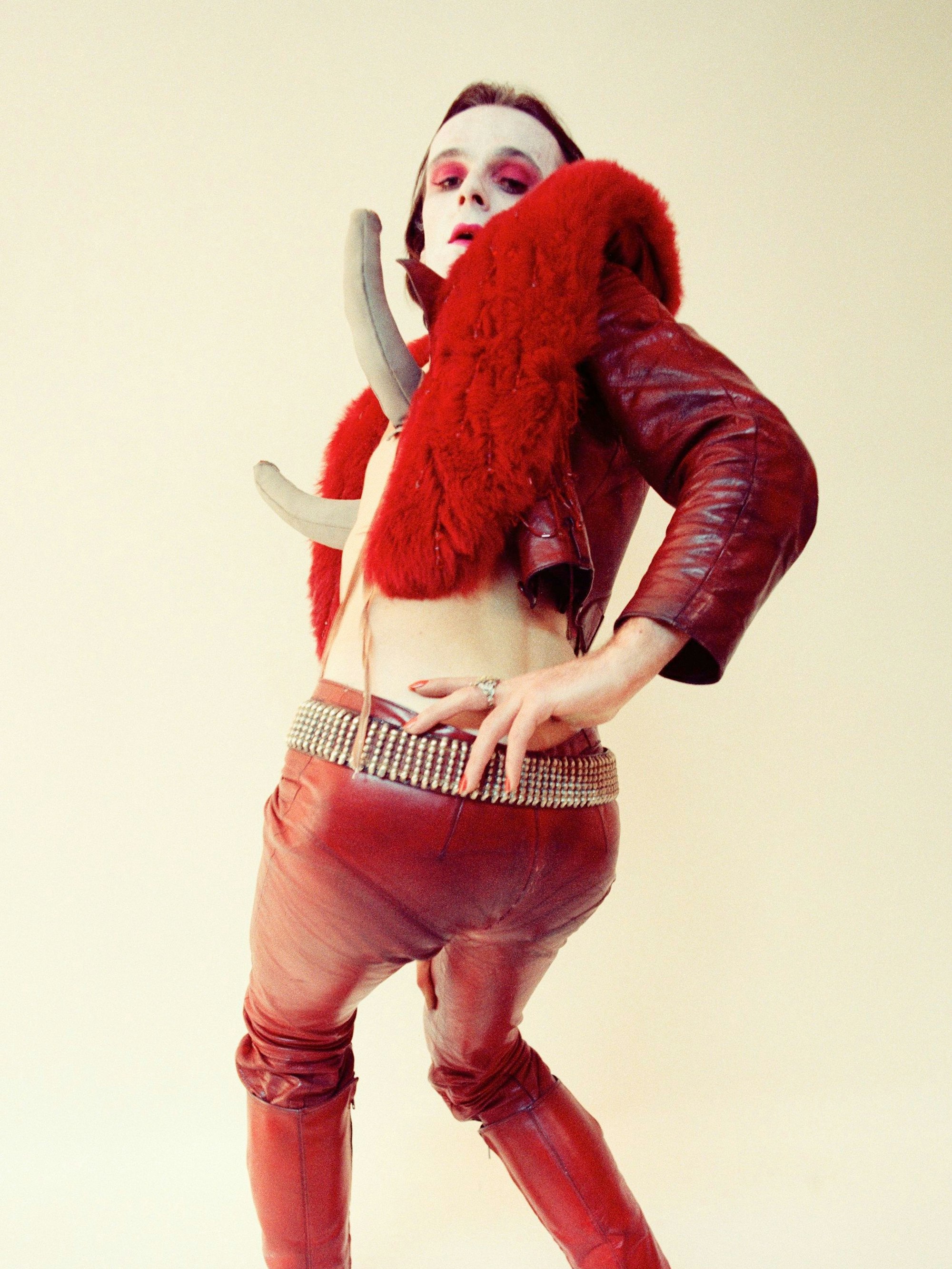 Jürgen Klauke posiert mit roter Lackhose, weiß geschminkten Gesicht und vor die Brust geschnallten Dildos.