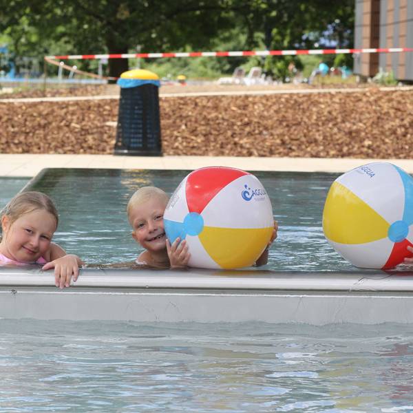 Drei Kinder plantschen mit Wasserbällen in einem Schwimmbecken.