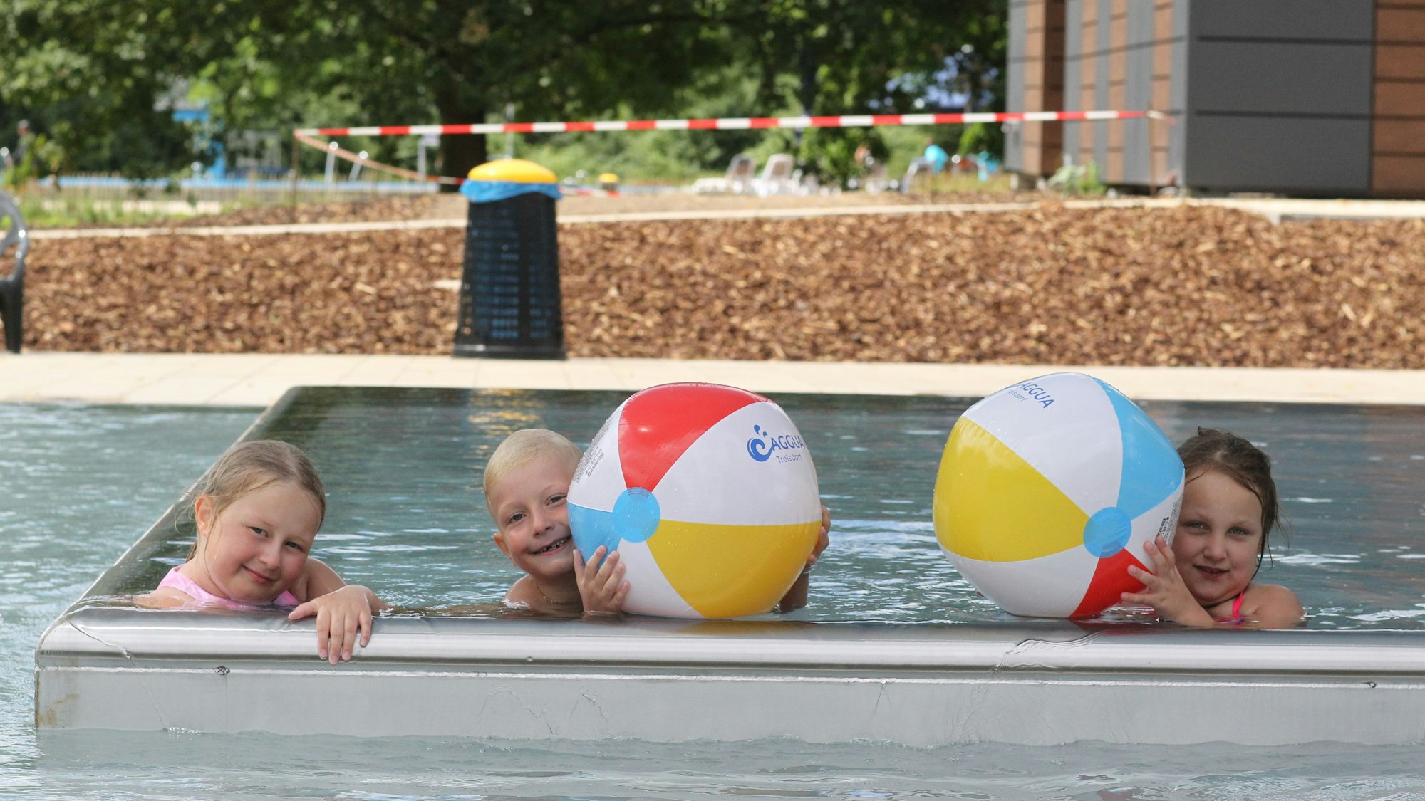 Drei Kinder plantschen mit Wasserbällen in einem Schwimmbecken.