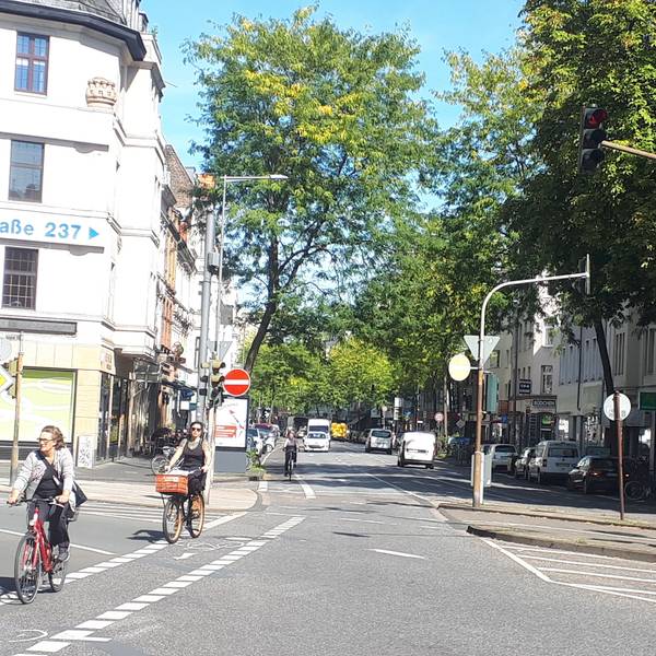 Die Kreuzung Neusser/Kempener/ Auerstraße ist zu sehen. Drei Straßen treffen aufeinander, Radfahrerinnen radeln über die Kreuzung.