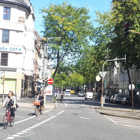 Die Kreuzung Neusser/Kempener/ Auerstraße ist zu sehen. Drei Straßen treffen aufeinander, Radfahrerinnen radeln über die Kreuzung.