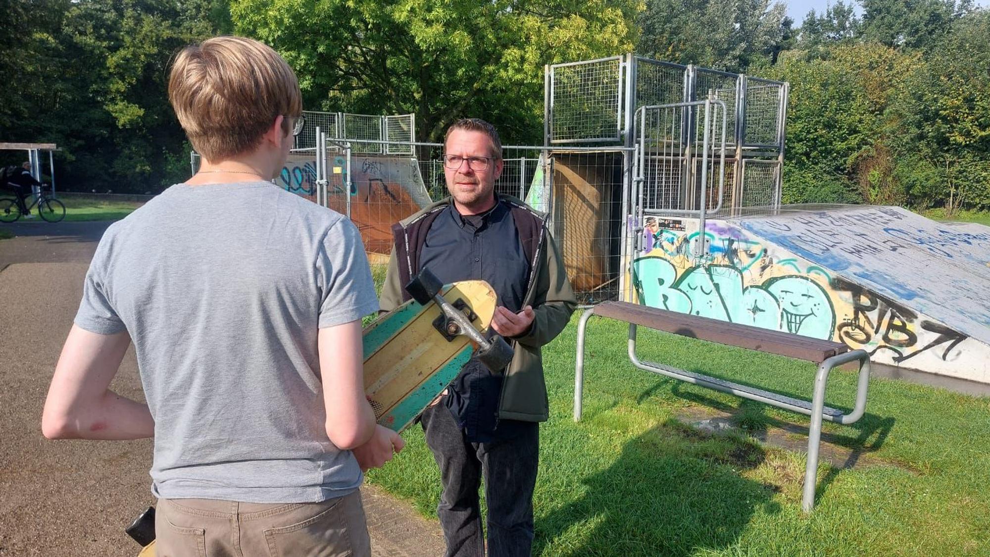 Ein Jugendlicher mit Skateboard in den Händen unterhält sich mit einem Mann vor einer Skateanlage.