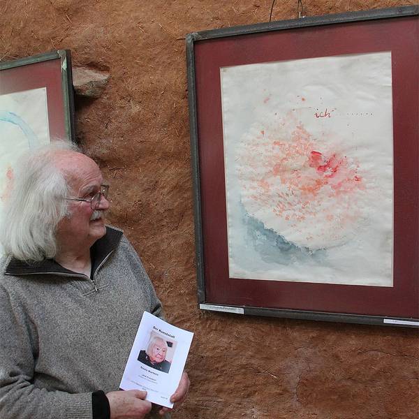 Wolfgang Martens steht an einem Gemälde, das an einer rot-braunen Wand hängt. Der Mann hat weißes Haar und trägt einen grauen Pullover.