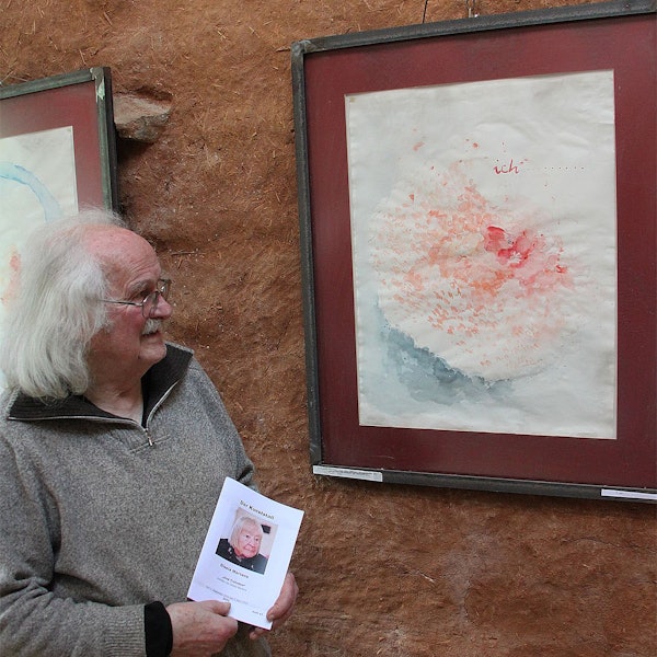 Wolfgang Martens steht an einem Gemälde, das an einer rot-braunen Wand hängt. Der Mann hat weißes Haar und trägt einen grauen Pullover.