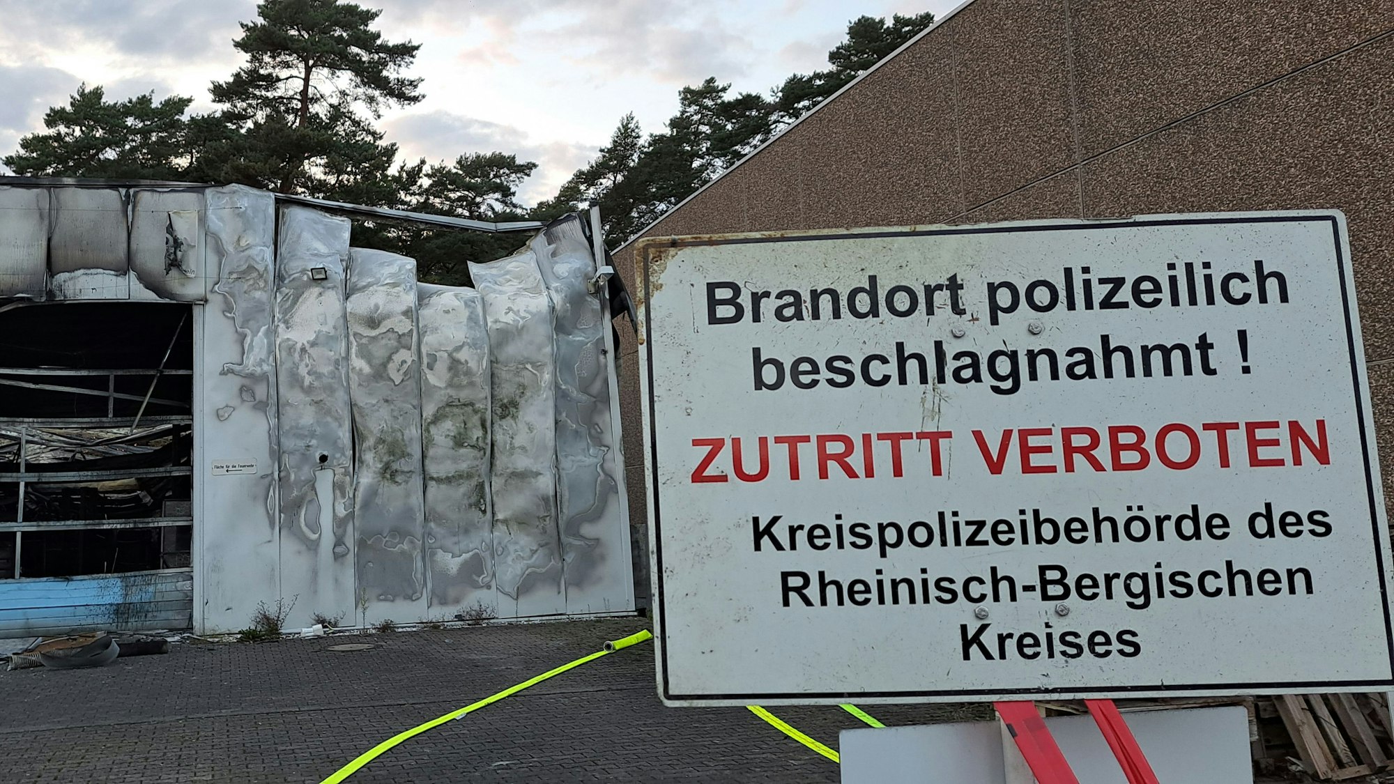 Ein Schild mit der Aufschrift „Brandort polizeilich beschlagnahmt“ steht vor einer ausgebrannten Gewerbehalle.