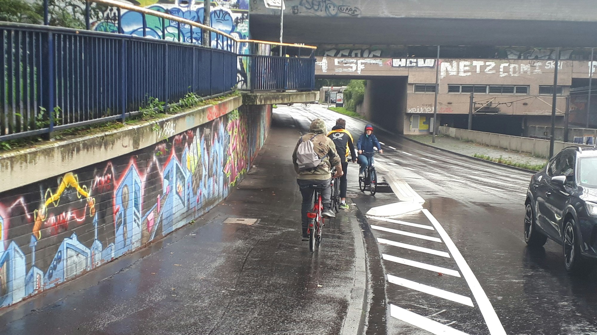 Auf einem Radweg kommen sich Radler entgegen. Links ist der Aufgang zur S-Bahn, rechts eine zweispurige Straße.