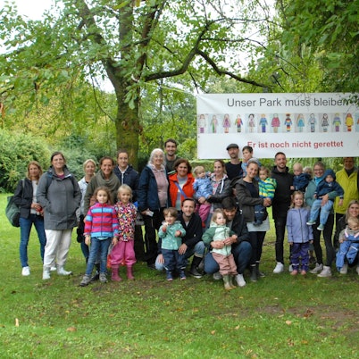 Männer, Frauen und Kinder stehen vor einem Banner, das in einem Park zwischen zwei Bäumen hängt.