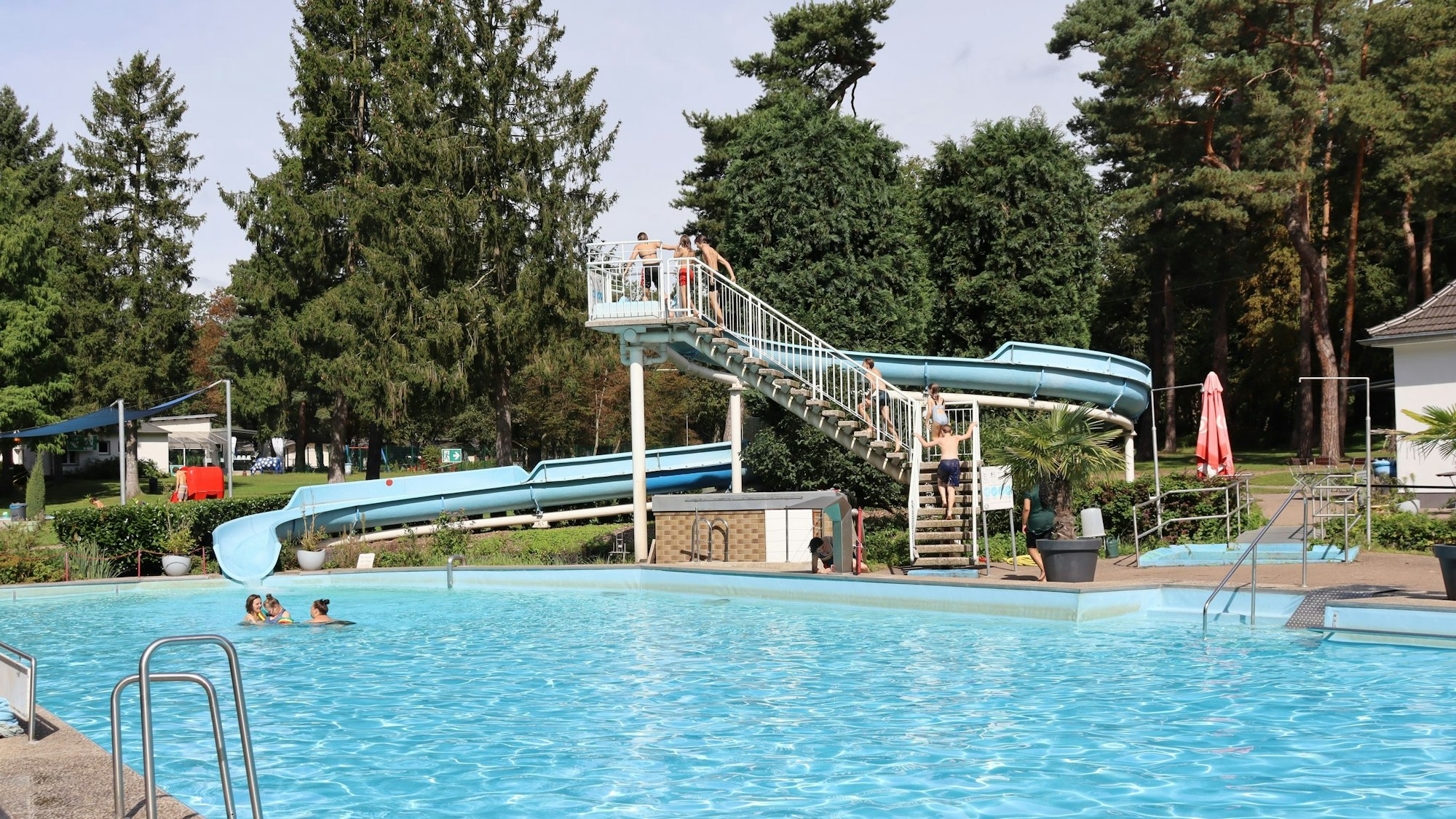Ein leicht besuchtes Freibad mit Wasserrutsche, im Hintergrund sind Bäume zu sehen.