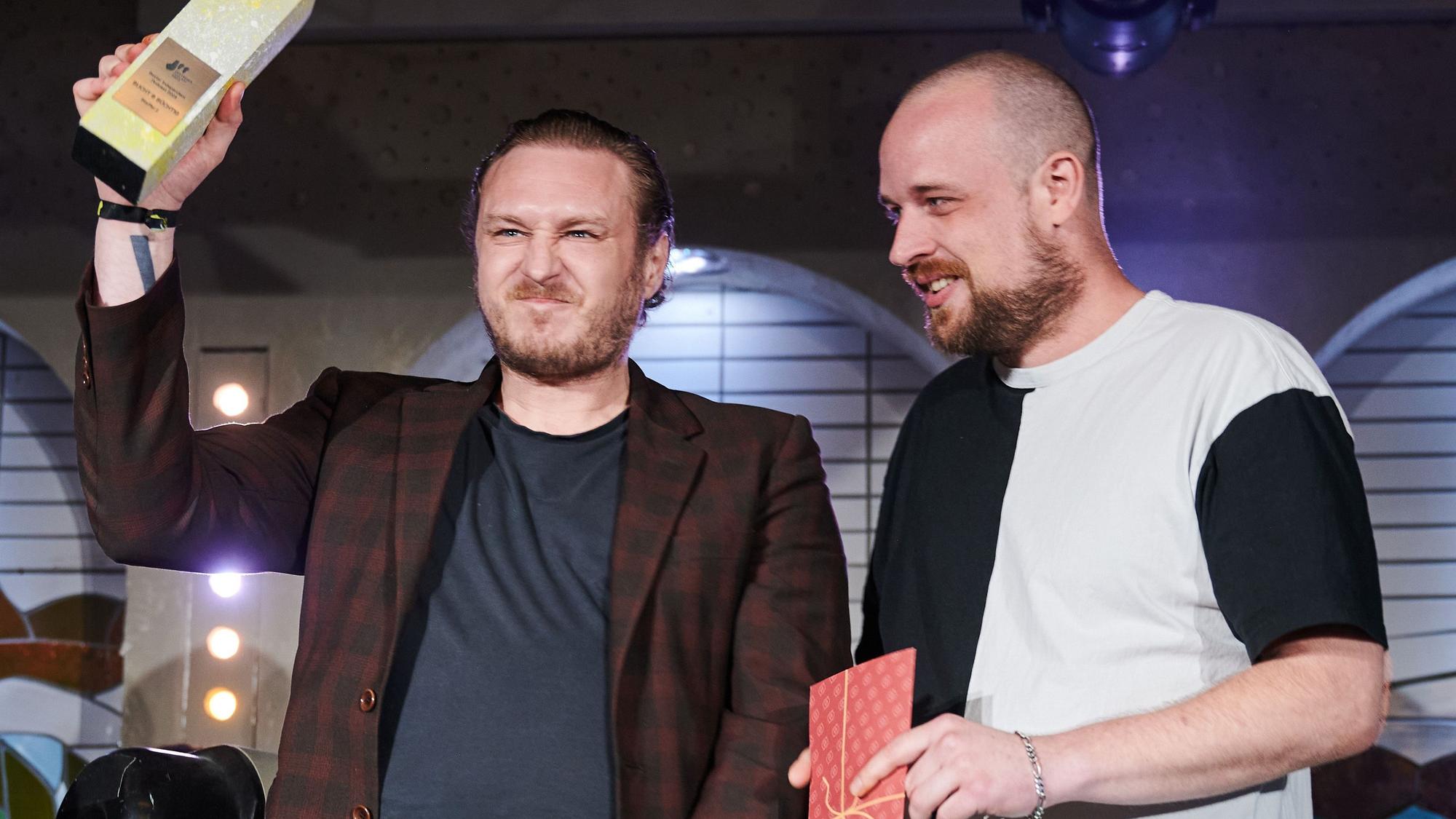 Decker hält den Award hoch und kneift die Augen zusammen, er trägt ein braun-kariertes Sakko. Cook hat einen kleinen roten Umschlag in der Hand und lächelt. Sein T-Shirt ist zur Hälfte schwarz, zur Hälfte weiß.