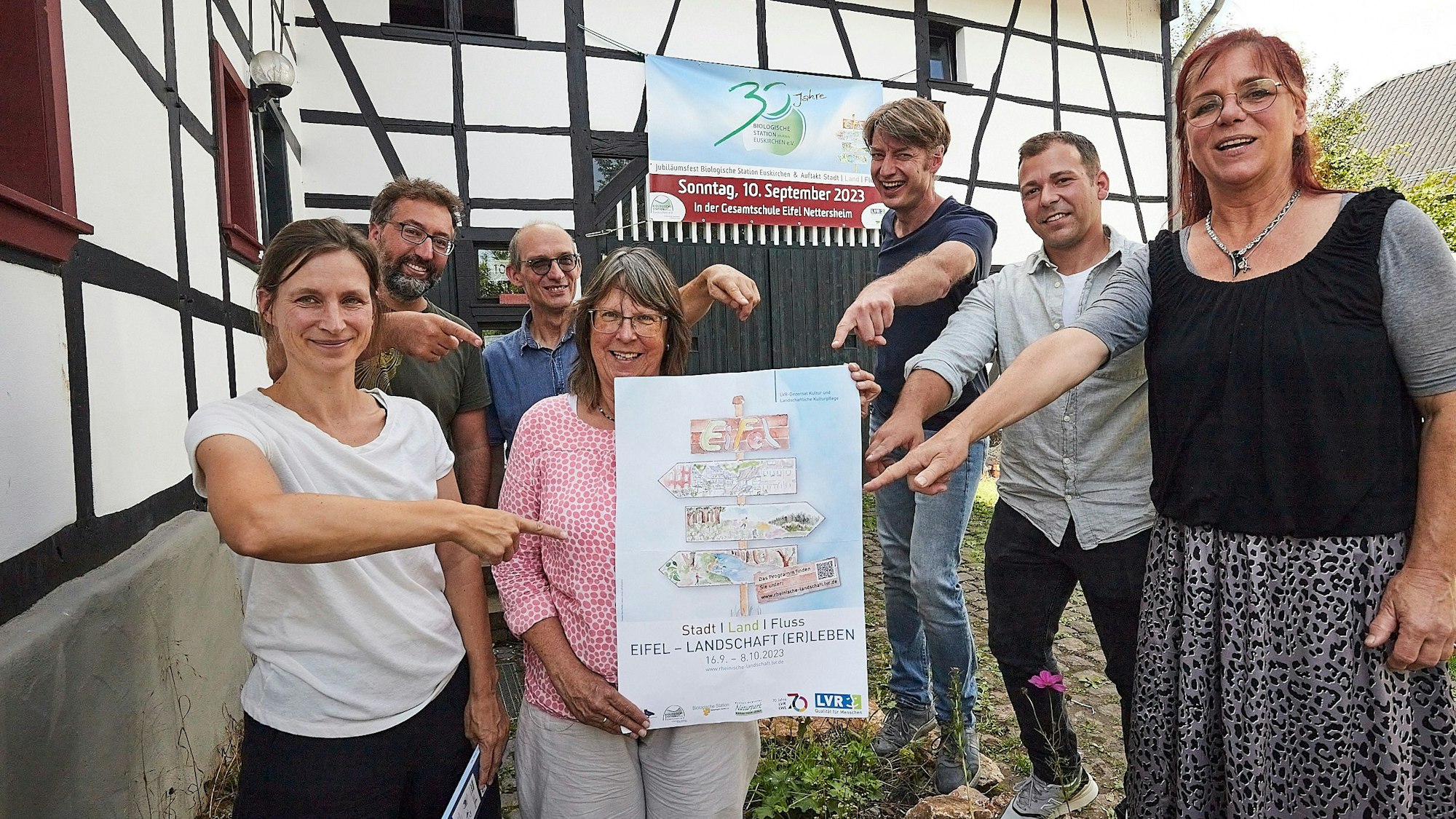 Einige Männer und Frauen stehen vor einem alten Fachwerkhaus. Sie zeigen auf ein Veranstaltungs-Plakat, das eine Frau in den Händen hält.