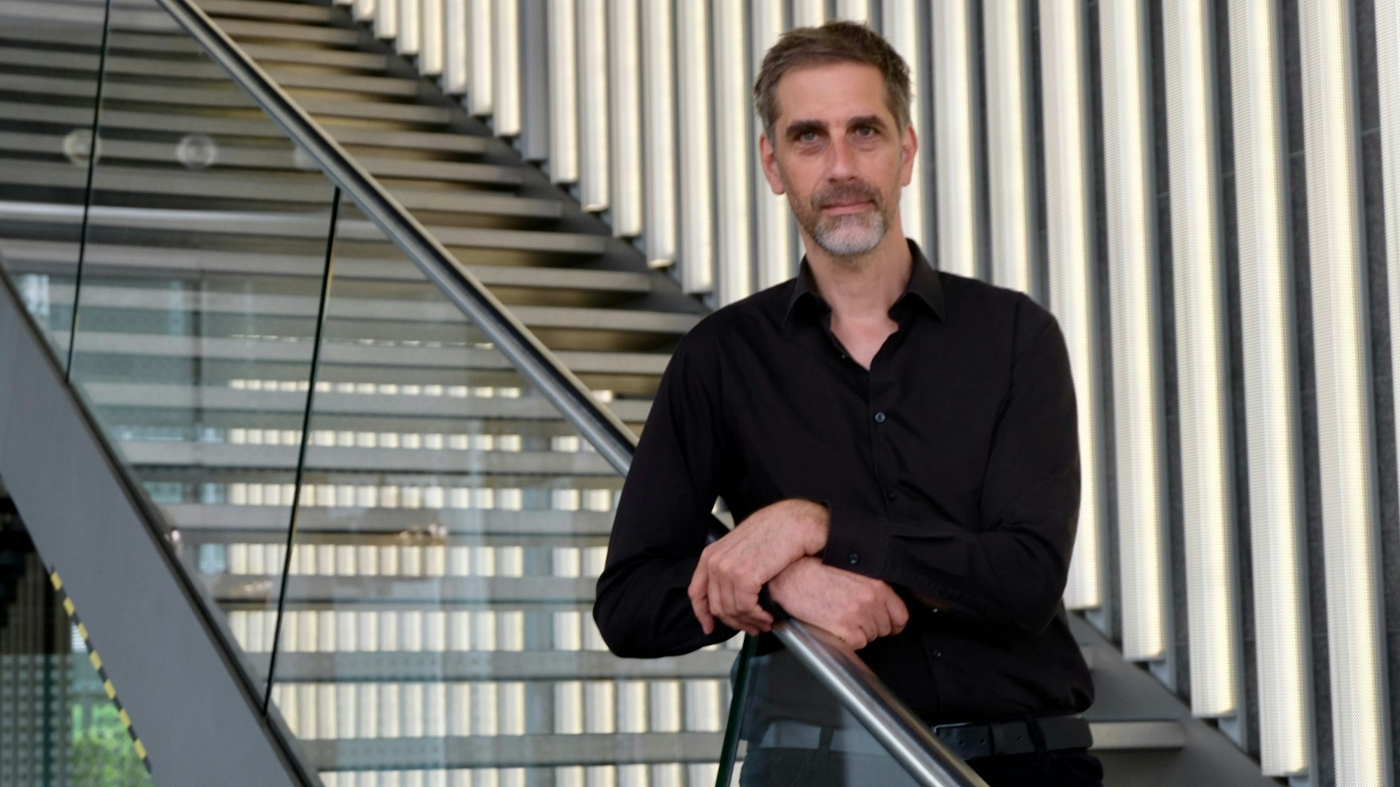 Ein Mann mit grauem Haar und Bart steht vor einem Fenster, im Hintergrund ist der Dom zu sehen.