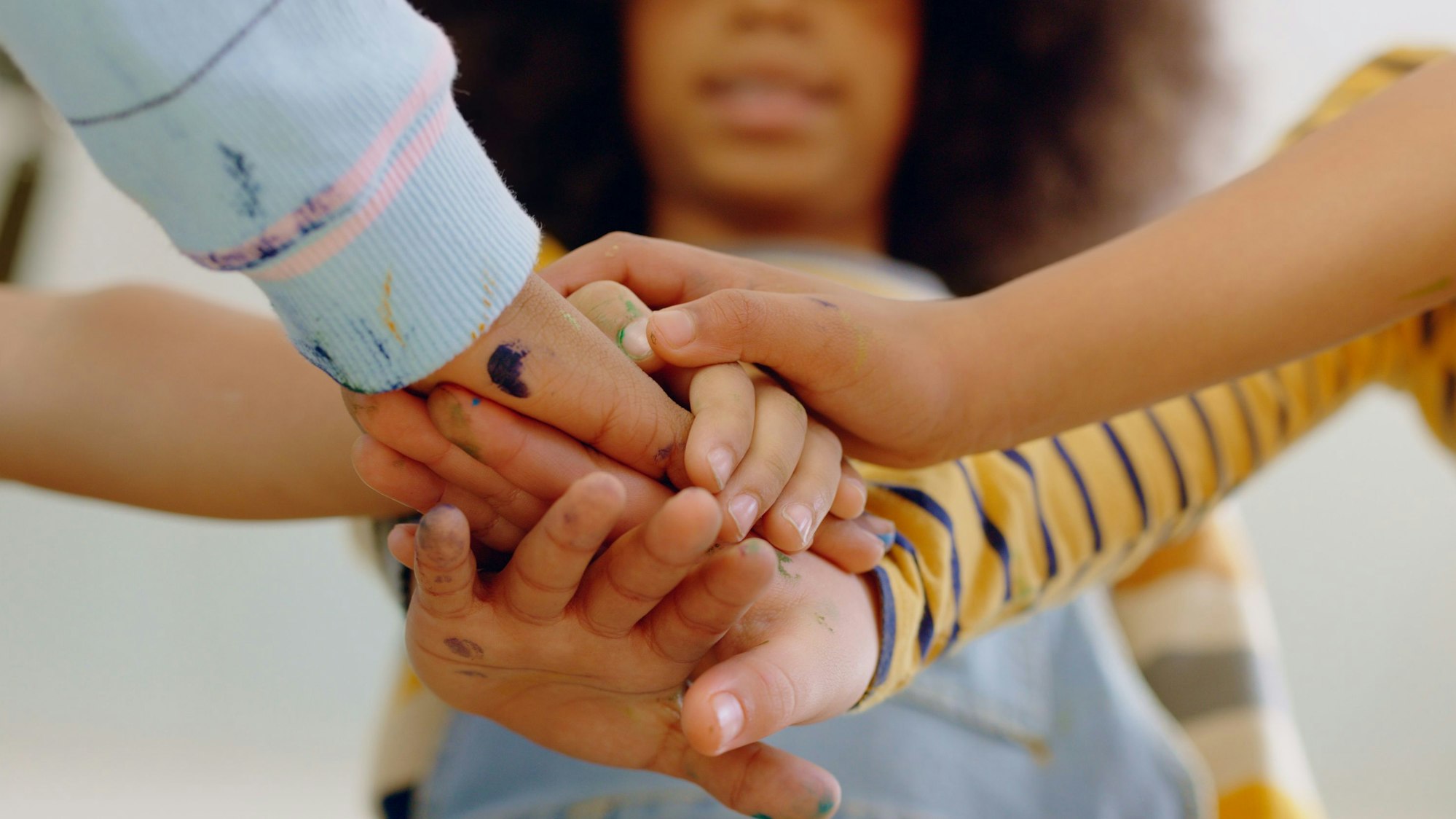Mehrere Kinderhände überkreuzen sich, im Hintergrund ist ein Mädchen mit dunkler Hautfarbe zu sehen.
