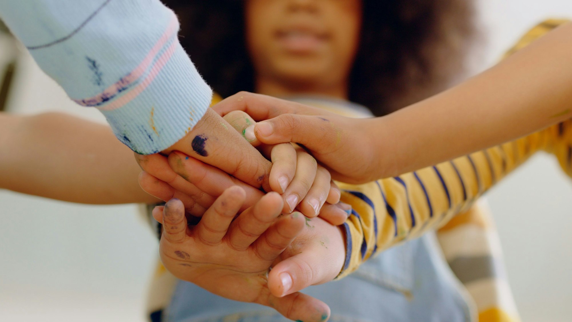 Mehrere Kinderhände überkreuzen sich, im Hintergrund ist ein Mädchen mit dunkler Hautfarbe zu sehen.