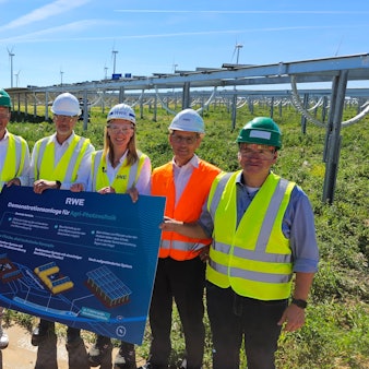 Fünf Männer und eine Frau in Warnwesten und mit Schutzbrillen und Helmen stehen vor einem Acker, auf dem Photovoltaikanlagen errichtet wurden.