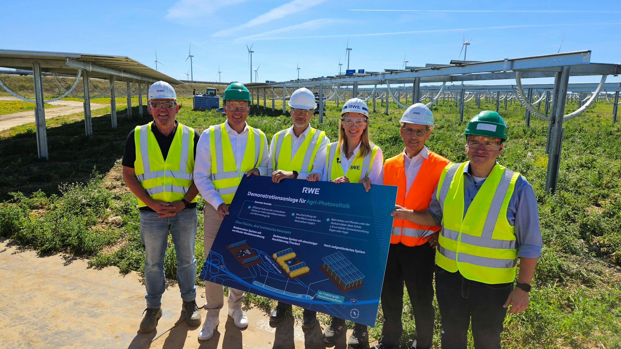 Fünf Männer und eine Frau in Warnwesten und mit Schutzbrillen und Helmen stehen vor einem Acker, auf dem Photovoltaikanlagen errichtet wurden.