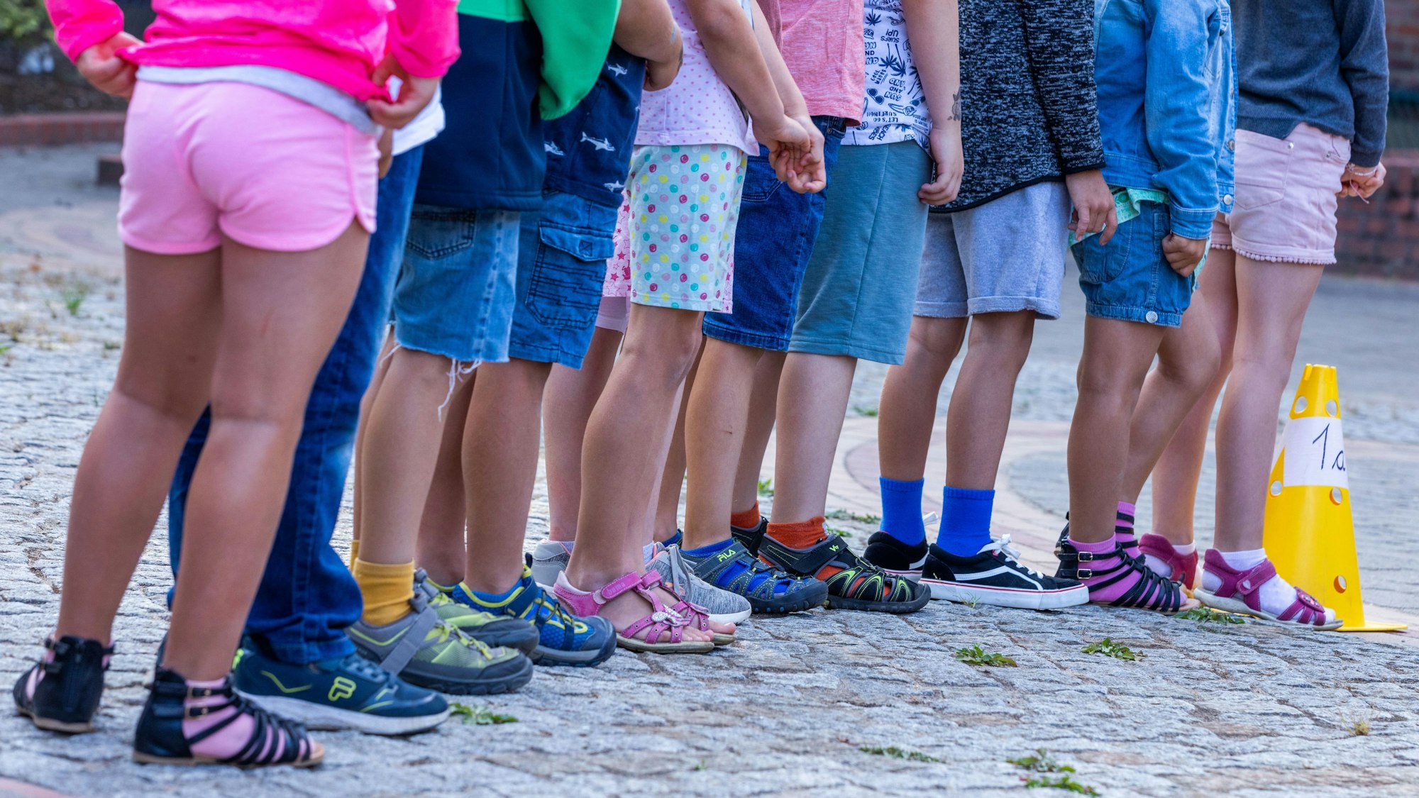 Schüler und Schülerinnen stehen in einer Reihe auf dem Schulhof einer Grundschule. Nur ihre Beine und die unterschiedlichen Schuhe sind zu sehen.