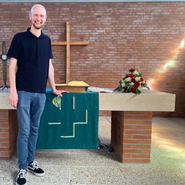 Diakon Jan Simons steht vor dem Altar der Evangelischen Kirche in Weilerswist. An der Wand hängt ein hölzernes Kreuz. Auf dem Altar liegen Blumen und die Bibel.