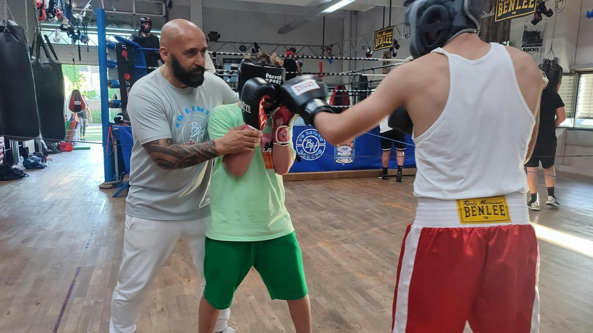 Bekim Hoxhaj zeigt zweien seiner Schützlinge die richtige Boxausrichtung. Im Hintergrund ist ein Boxring mit trainierenden Boxern zu sehen.
