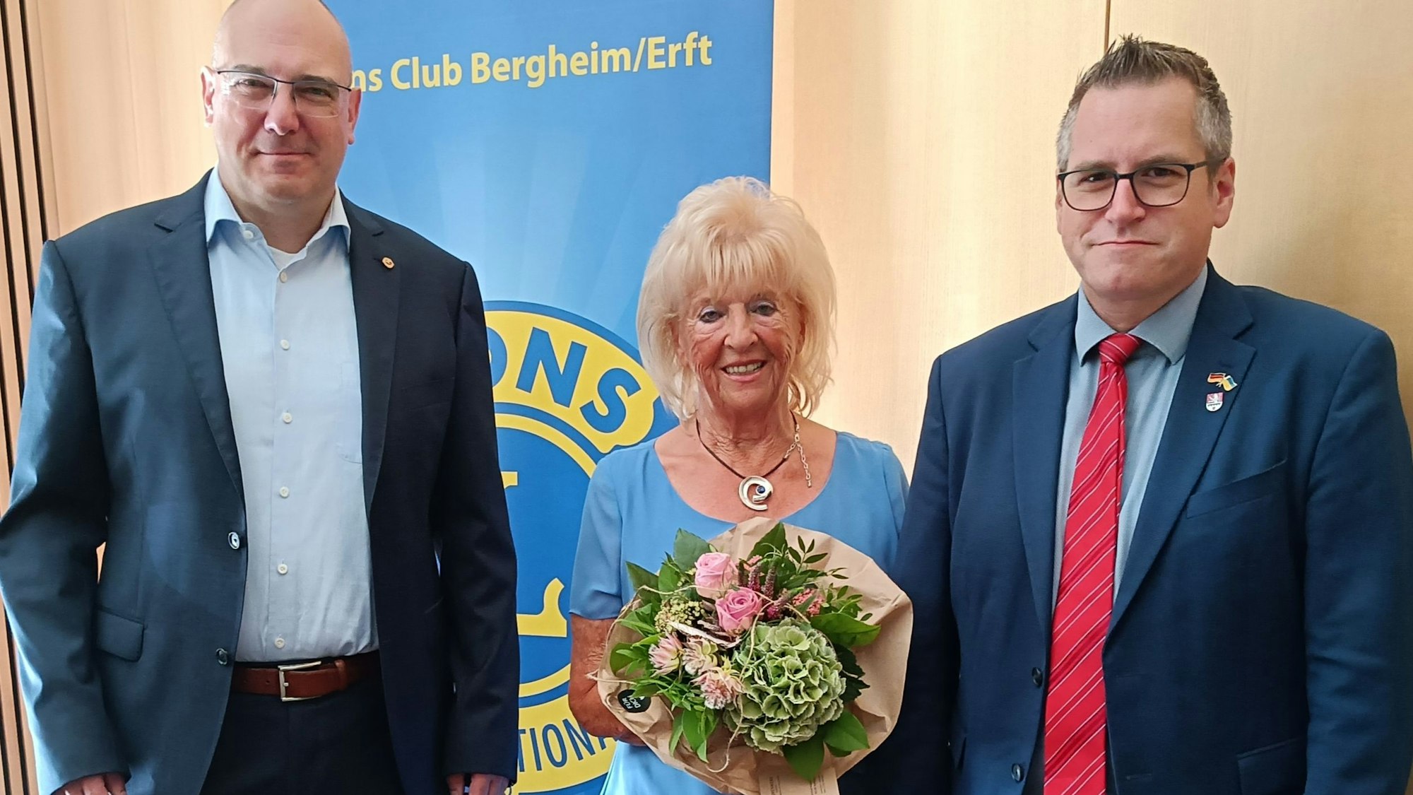 Eine Frau in einem blauem Kleid mit einem Blumenstrauß steht zwischen zwei Männern in Anzügen.