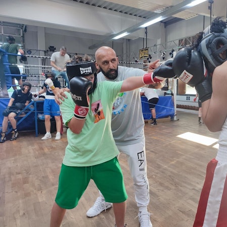 Box-Studiobesitzer Bekim Hoxhaj trainiert er zwei jüngere Kämpfer. Sie stehen sich mit Boxhandschuhen und Kopfschutz gegenüber. Ein Kämpfer zielt mit seinem Schlag auf Kopfhöhe des Gegenübers, der die Handschuhe schützend vor das Gesicht hält.