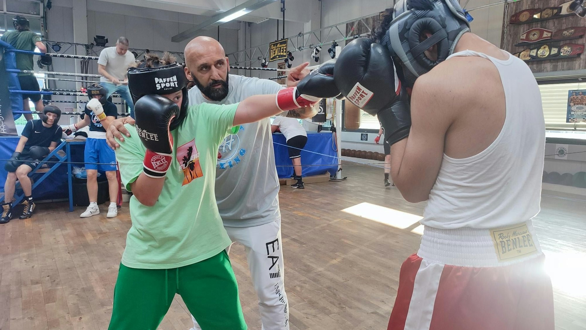 Box-Studiobesitzer Bekim Hoxhaj trainiert er zwei jüngere Kämpfer. Sie stehen sich mit Boxhandschuhen und Kopfschutz gegenüber. Ein Kämpfer zielt mit seinem Schlag auf Kopfhöhe des Gegenübers, der die Handschuhe schützend vor das Gesicht hält.