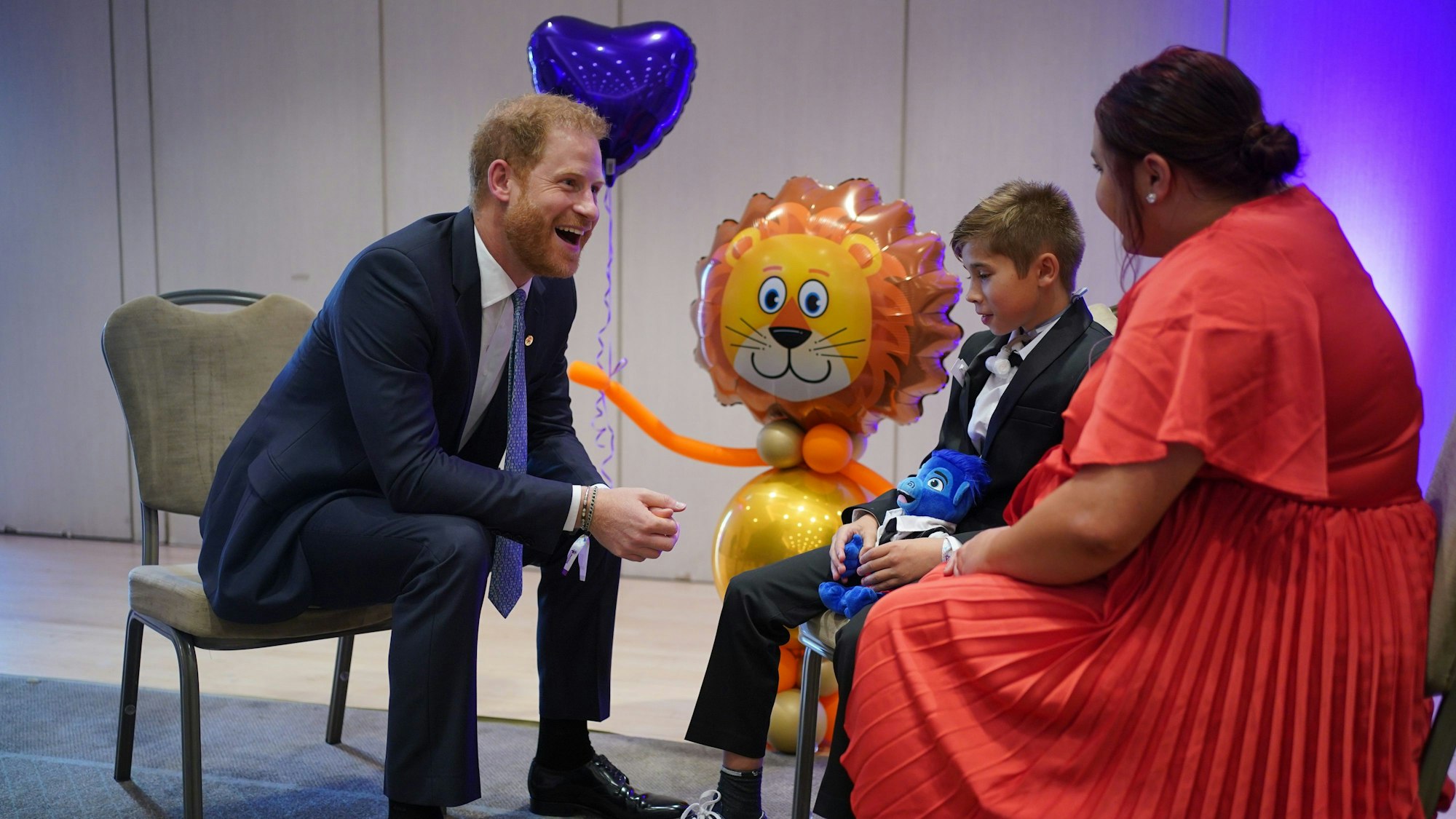 Prinz Harry grinst breit - ihm gegenüber sitzen ein Junge und eine Frau - im Hintergrund befinden sich Luftballons.