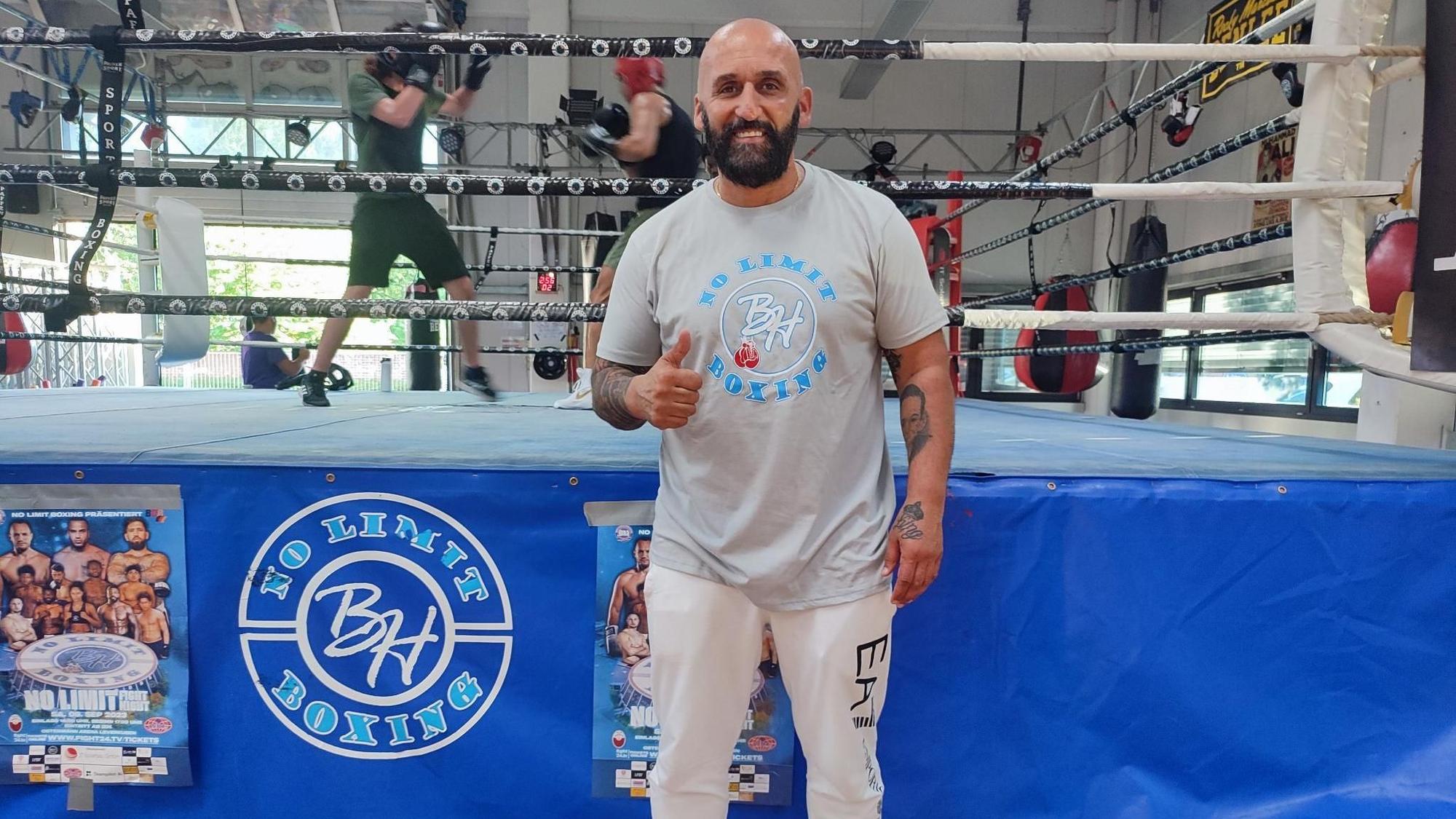 Boxen-Trainer, -Promoter und Studiobesitzer Bekim Hoxhaj steht in seinem Boxstudio vor einem Boxring und reckt einen Daumen hoch.