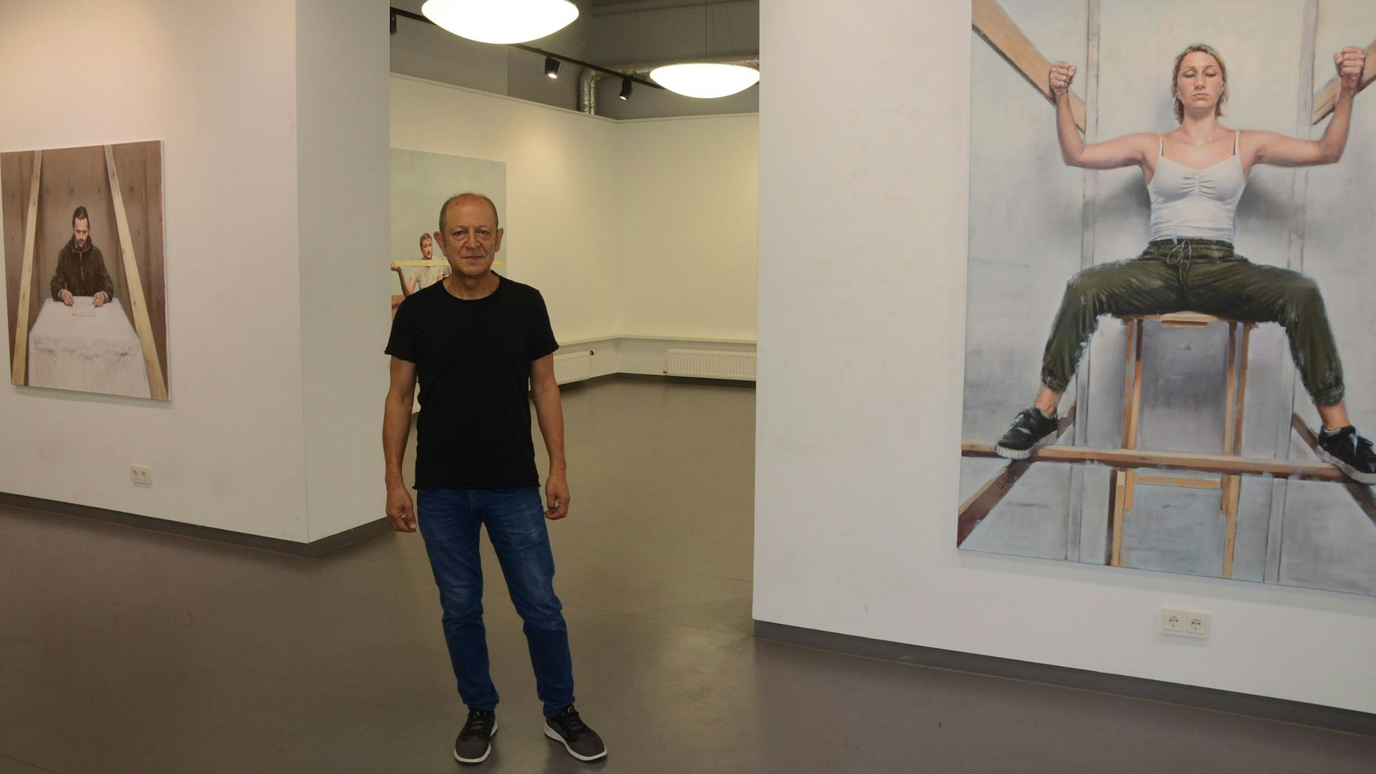 Ein Mann im schwarzen T-Shirt, Jeans und dunklen Sneakern steht im Ausstellungsraum. Links und rechts hängen Bilder, die er gemalt hat.