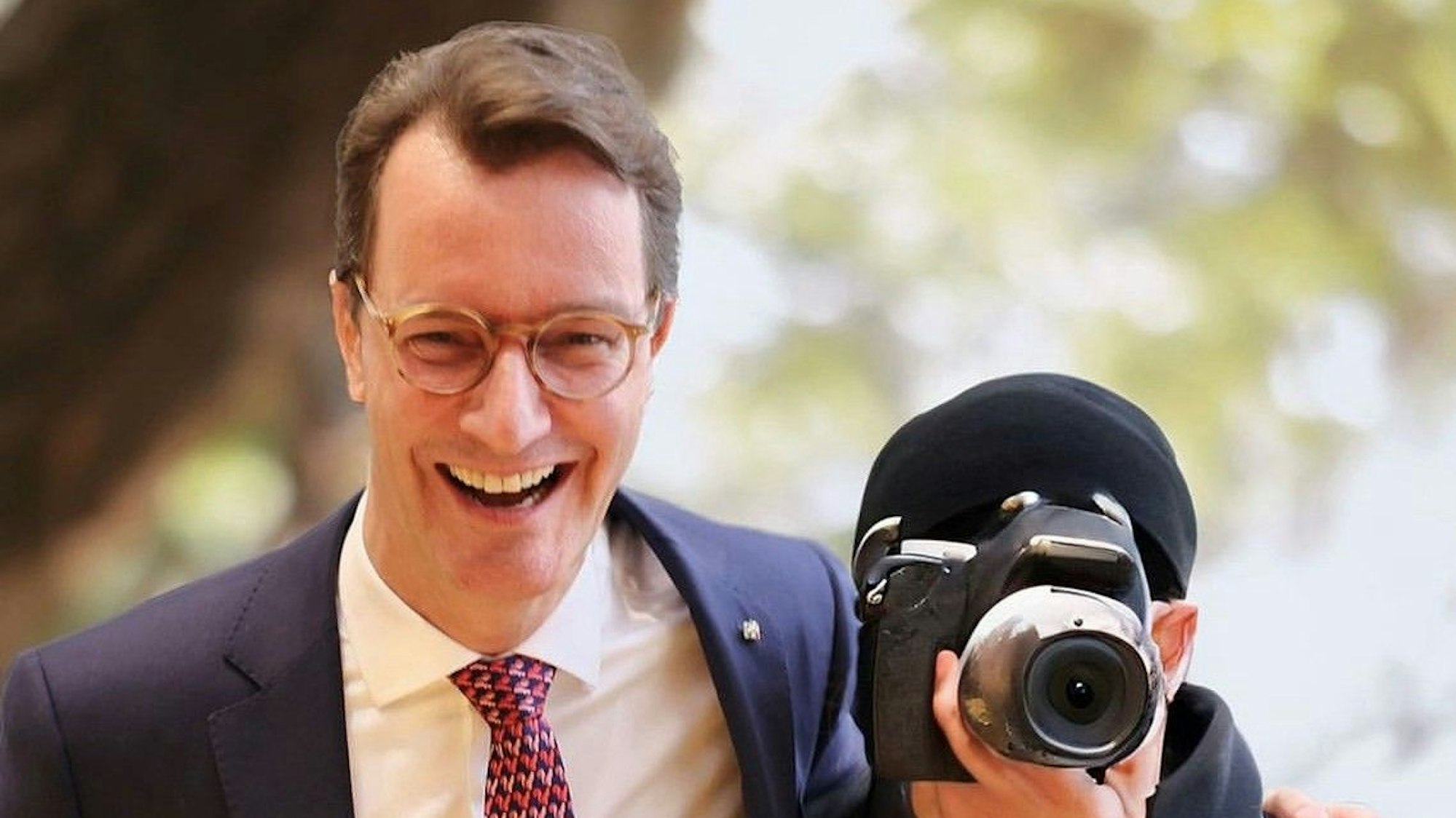 Auf einem gefälschten Bild ist Ministerpräsident Wüst zu sehen, wie er lachend einem Fotografen den Arm um die Schulter legt. Dessen Gesicht ist hinter einer Kamera verborge.