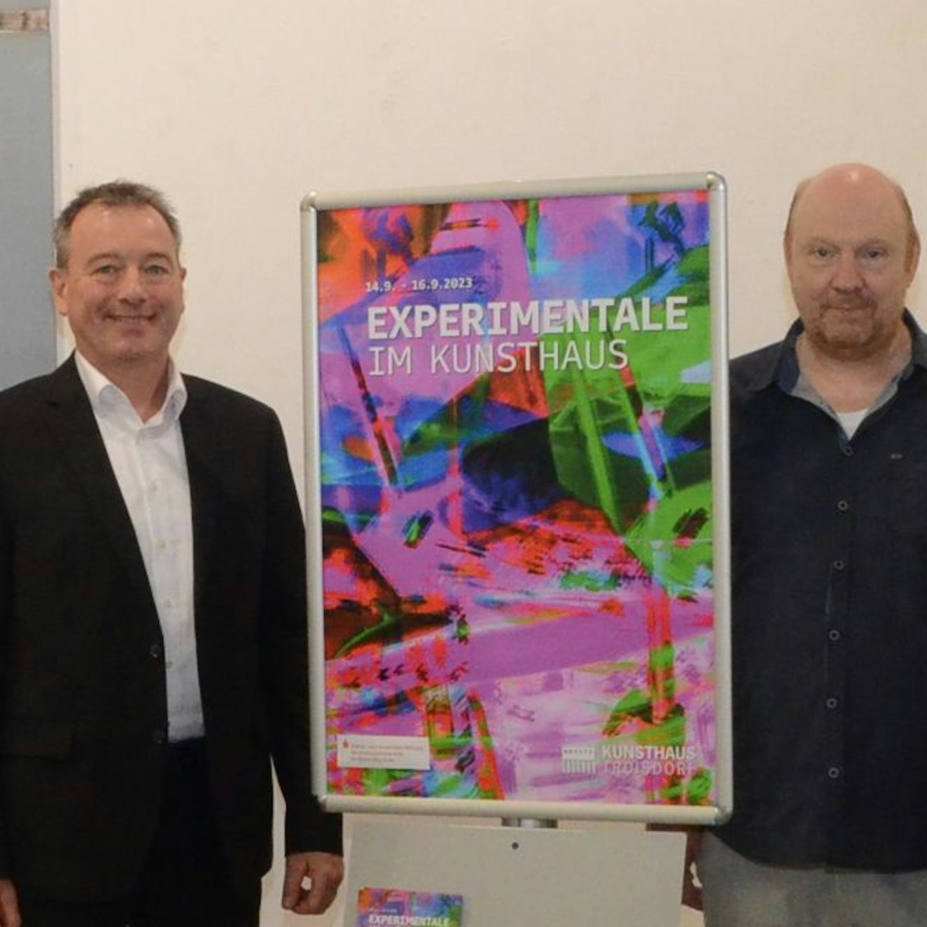 Ein Mann im Anzug und ein zweiter im dunklen Hemd stehen neben einem grellbunten Plakat. Es wirbt für die Experimentale im Kunsthaus.