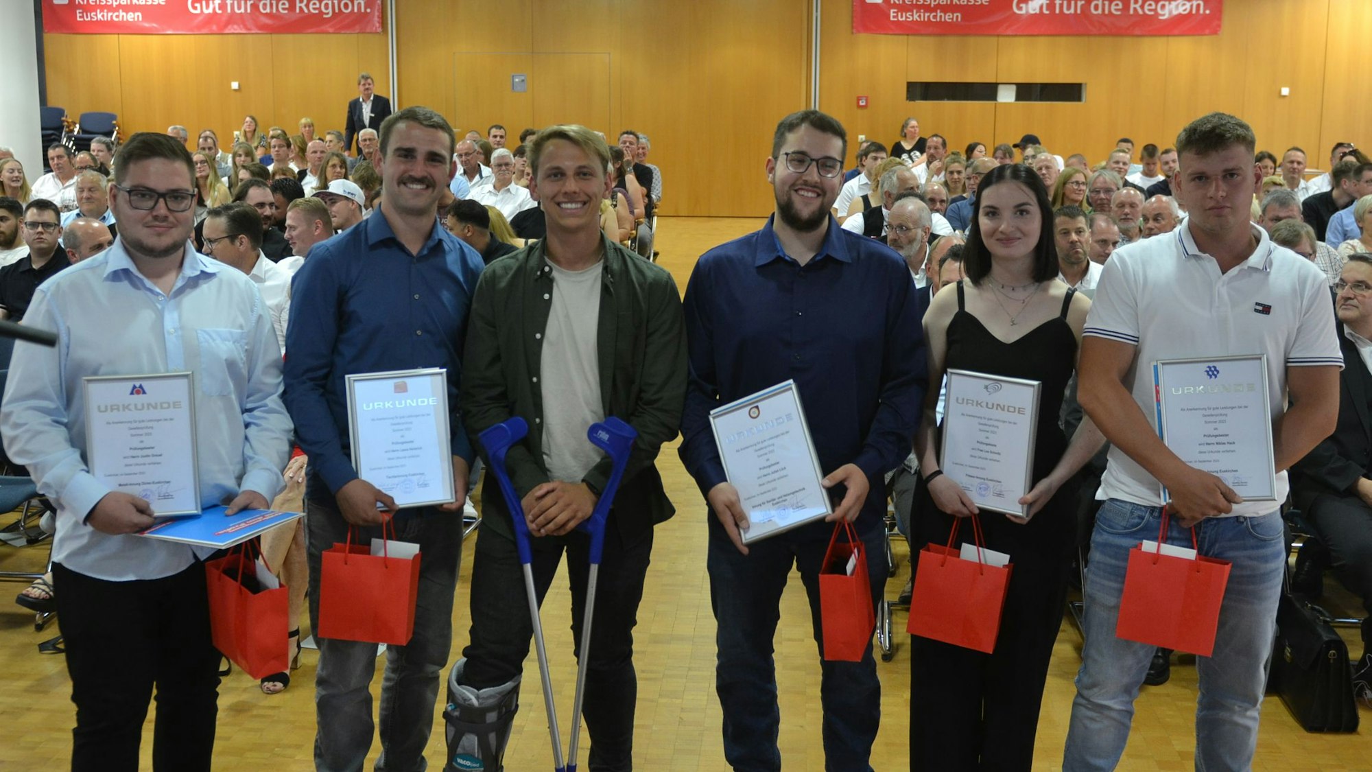 Fünf junge Männer und eine Frau stehen in einem großen Saal und halten eine Urkunde in den Händen. Im Hintergrund ist Publikum zu erkennen.