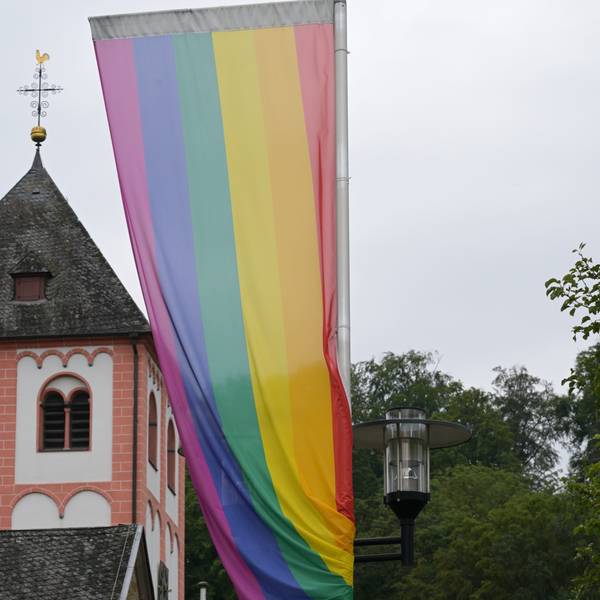 Eine Regenbogenflagge mit sechs Farbstreifen weht im Wind. Im Hintergrund ein Kirchturm.