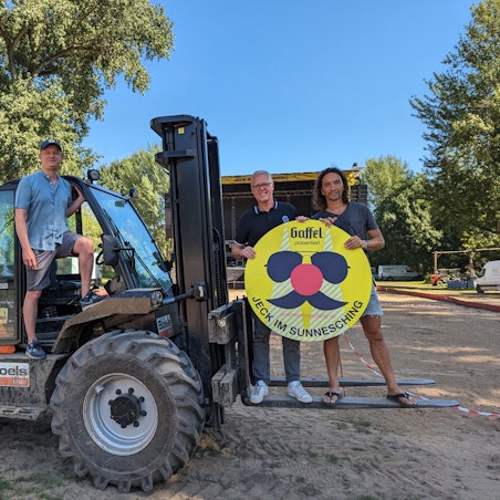 Die drei Männer stehen auf einem Gabelstapler. Deloy und Brings halten ein Schild von Jeck im Sunnesching hoch.
