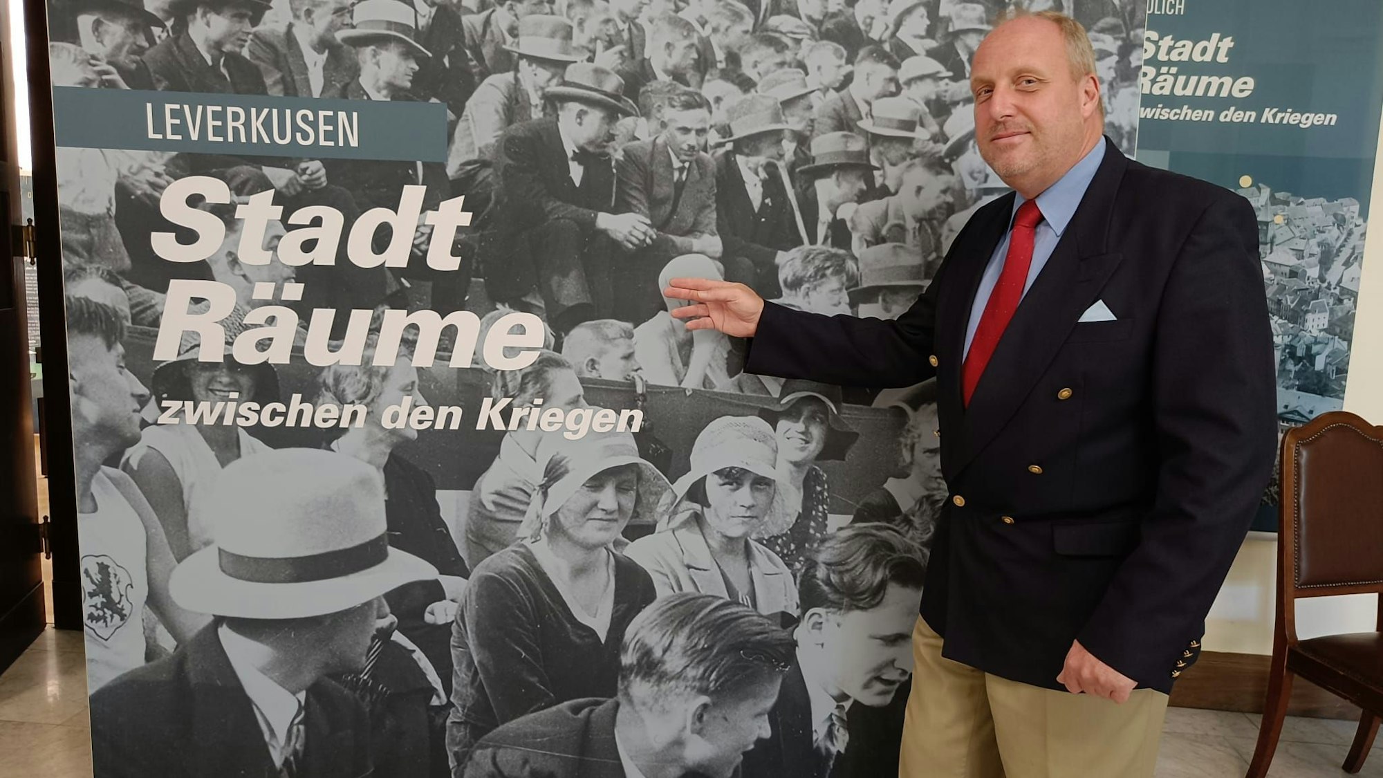 Ein Herr steht vor einer Wandtafel und weist mit der Hand auf sie. Darauf abgebildet ist ein historisches Foto, dass Menschen verschiedenen Alters auf einer Tribüne zeigt sowie der Schriftzug „Leverkusen. StadtRäume zwischen den Weltkriegen.“