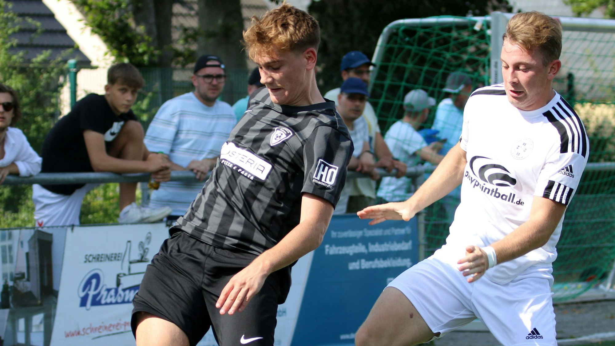 Zwei Fußballer im Kampf um den Ball, im Hintergrund erkennt man die Zuschauer hinter einer Bande.