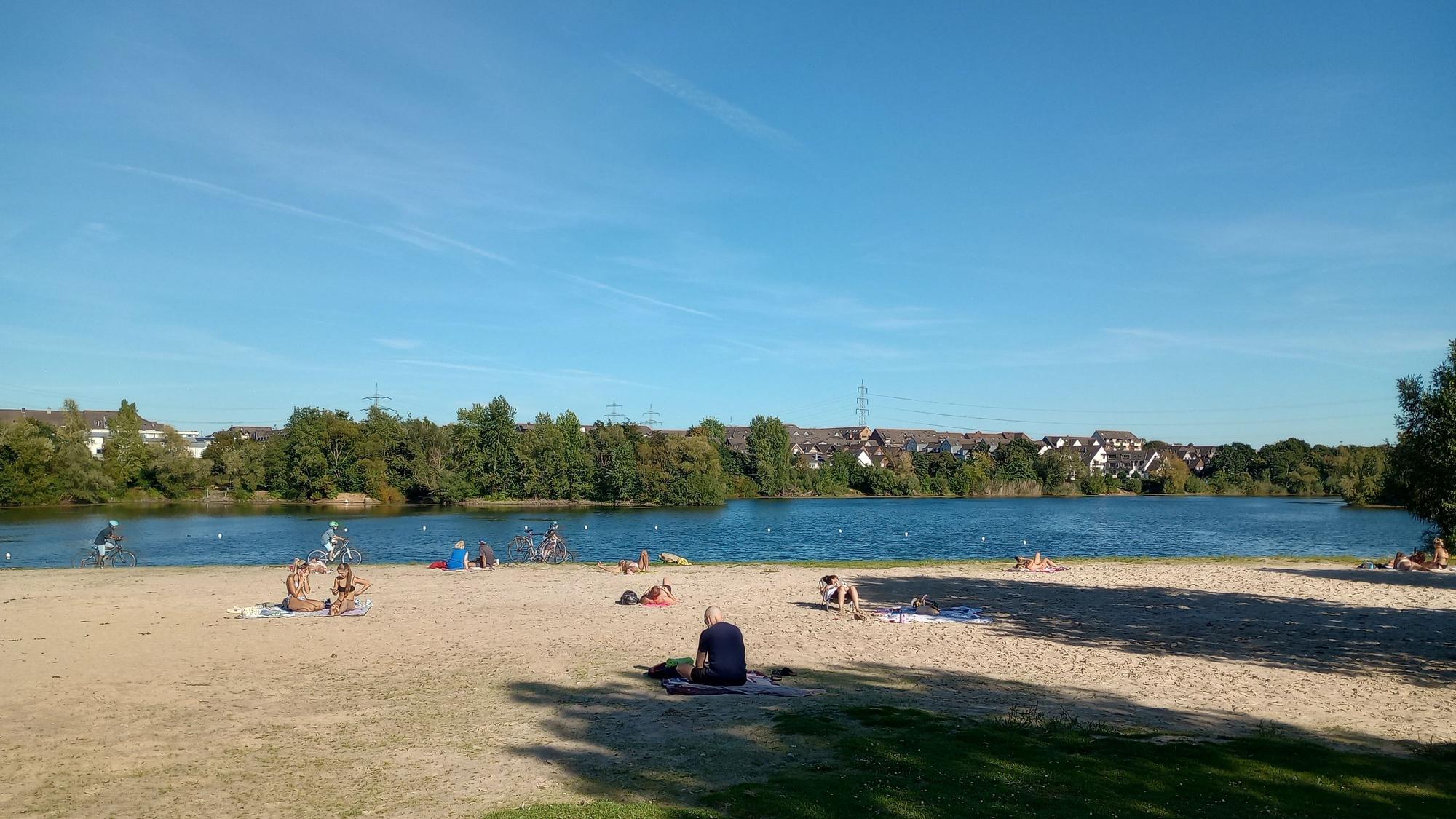 Menschen sitzen am sandigen Ufer eines Baggersees.