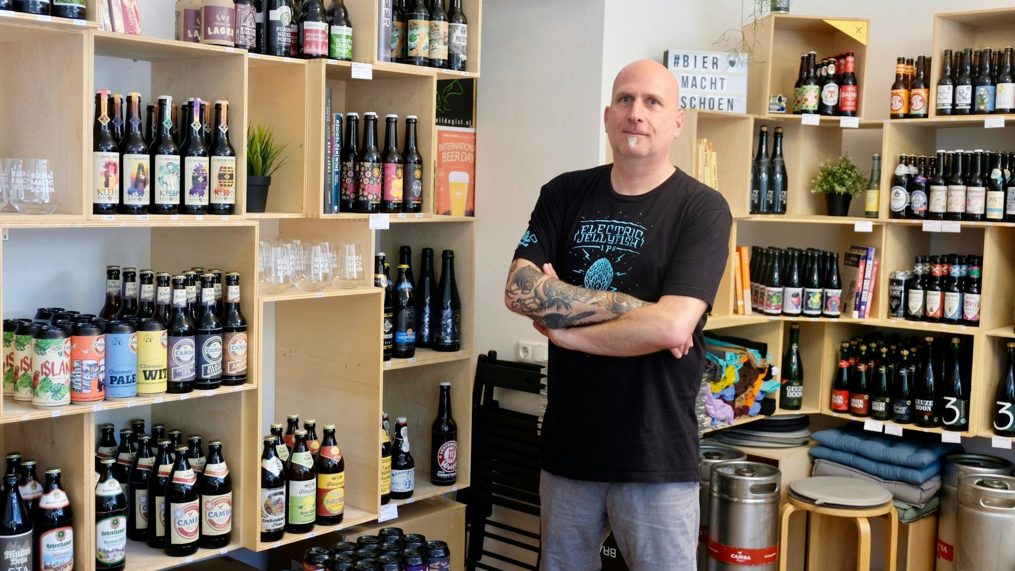 Ein Mann mit Glatzkopf und kleinem Ziegenbart steht in einem Laden, in dem die Regale vollgestellt sind mit Bierflaschen.