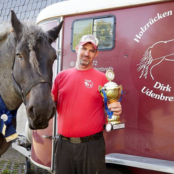 Dirk Zöll hält einen Pokal in der Hand. Mit seinem Belgischen Kaltblut Wotan steht er am Transportanhänger, den als Logo ein Holzrückepferd zeigt.