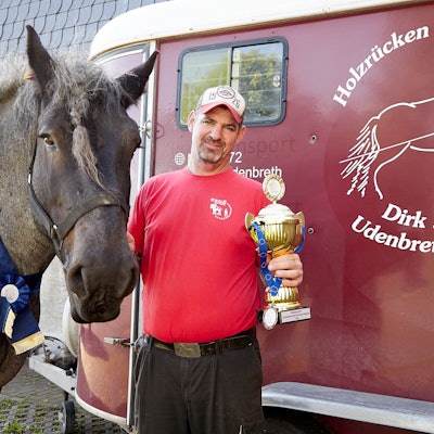 Dirk Zöll hält einen Pokal in der Hand. Mit seinem Belgischen Kaltblut Wotan steht er am Transportanhänger, den als Logo ein Holzrückepferd zeigt.