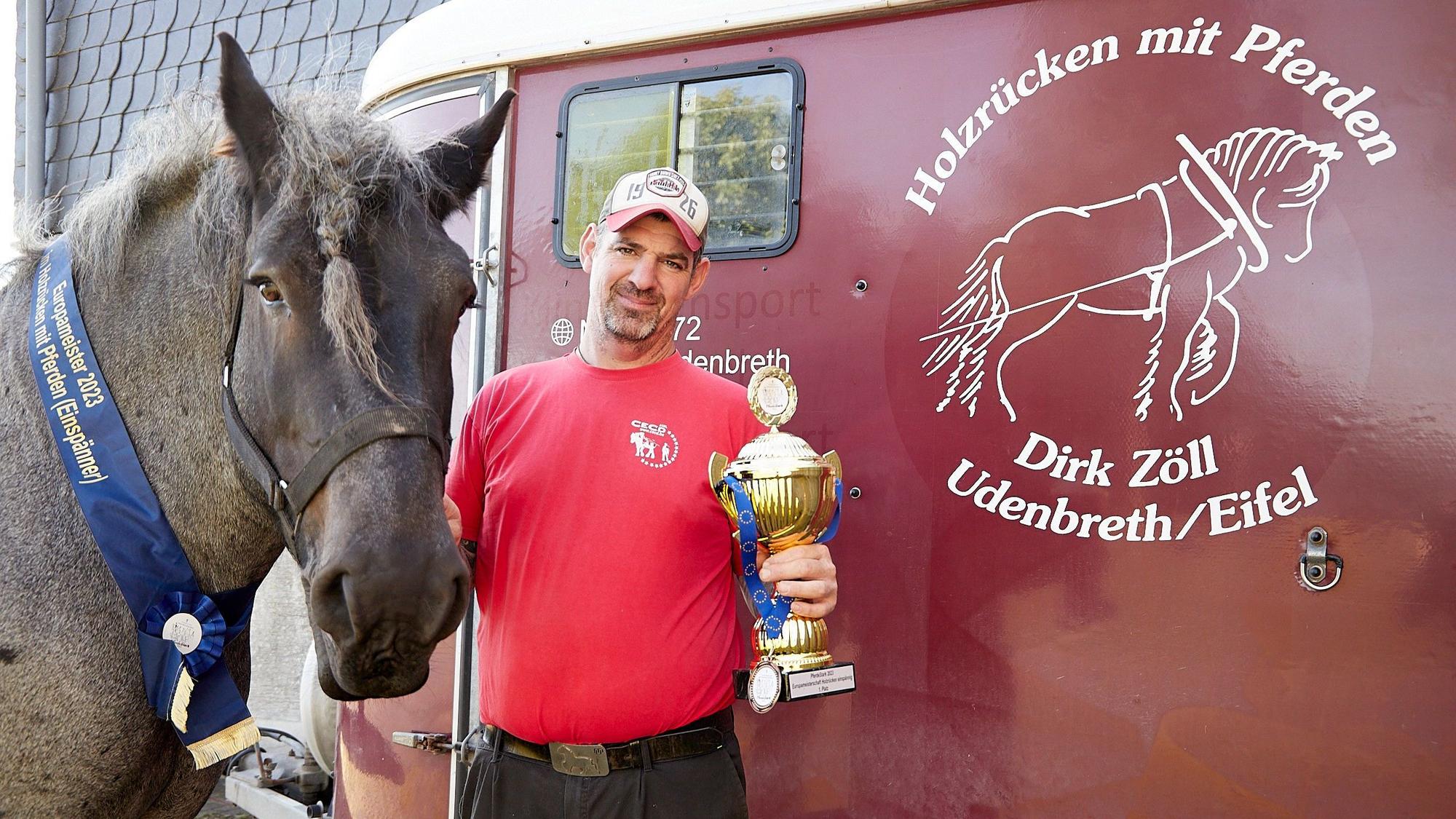Dirk Zöll hält einen Pokal in der Hand. Mit seinem Belgischen Kaltblut Wotan steht er am Transportanhänger, den als Logo ein Holzrückepferd zeigt.