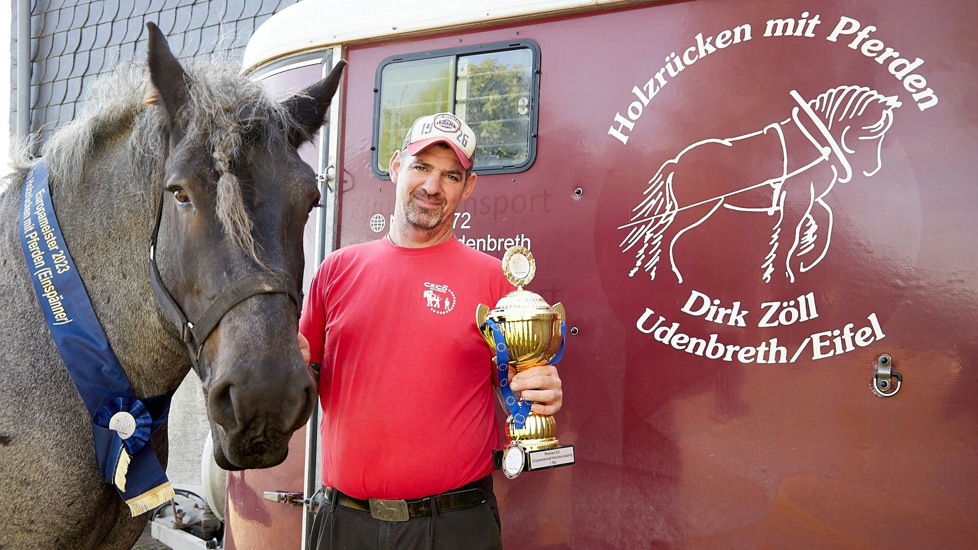Dirk Zöll hält einen Pokal in der Hand. Mit seinem Belgischen Kaltblut Wotan steht er am Transportanhänger, den als Logo ein Holzrückepferd zeigt.