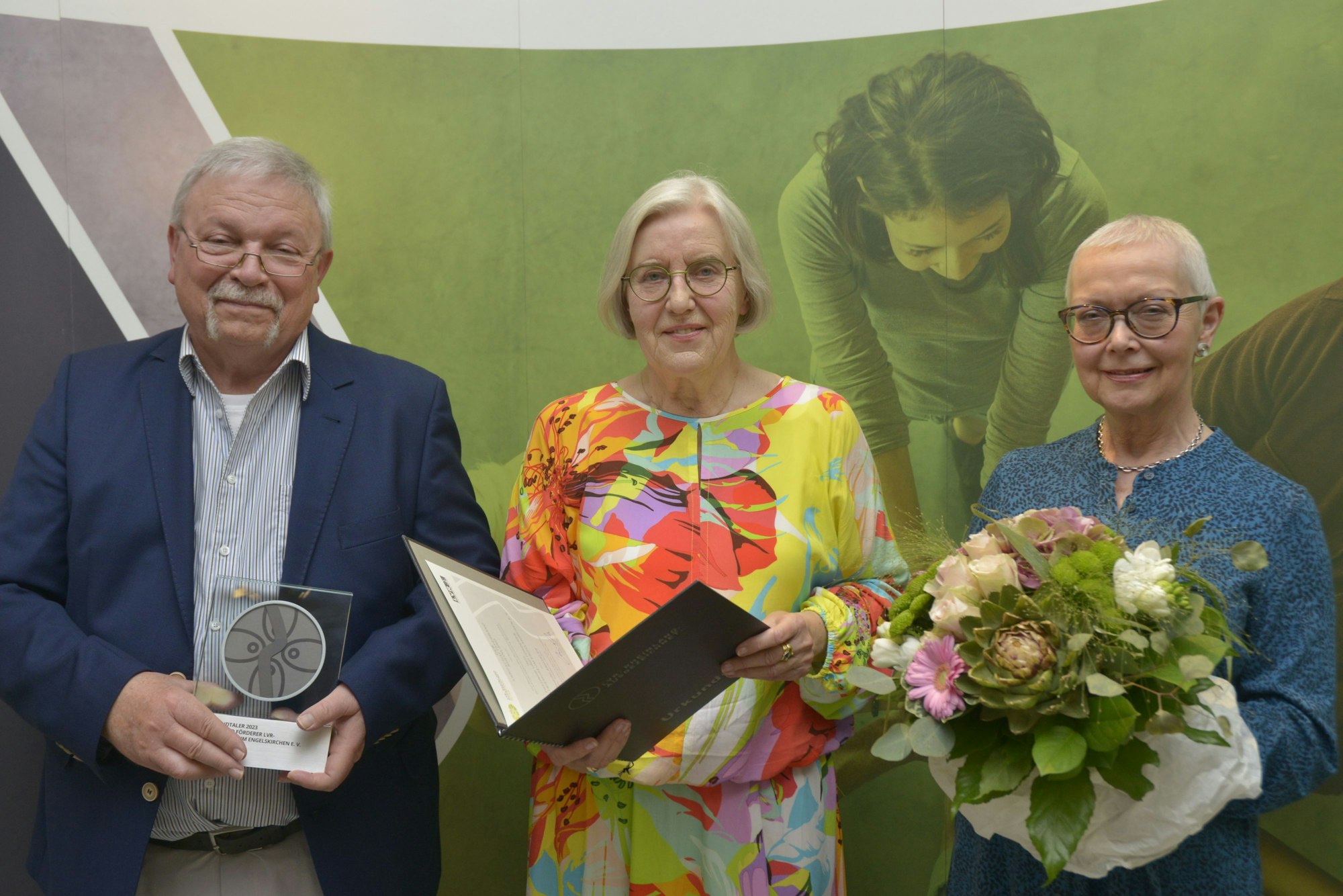 Das Foto zeigt von links nach rechts Peter Ruland mit dem Rheinlandtaler, Ursula Mahler, die stellvertretende Vorsitzende der Landschaftsversammlung Rheinland und Maria Clever mit einem Blumenstrauß.