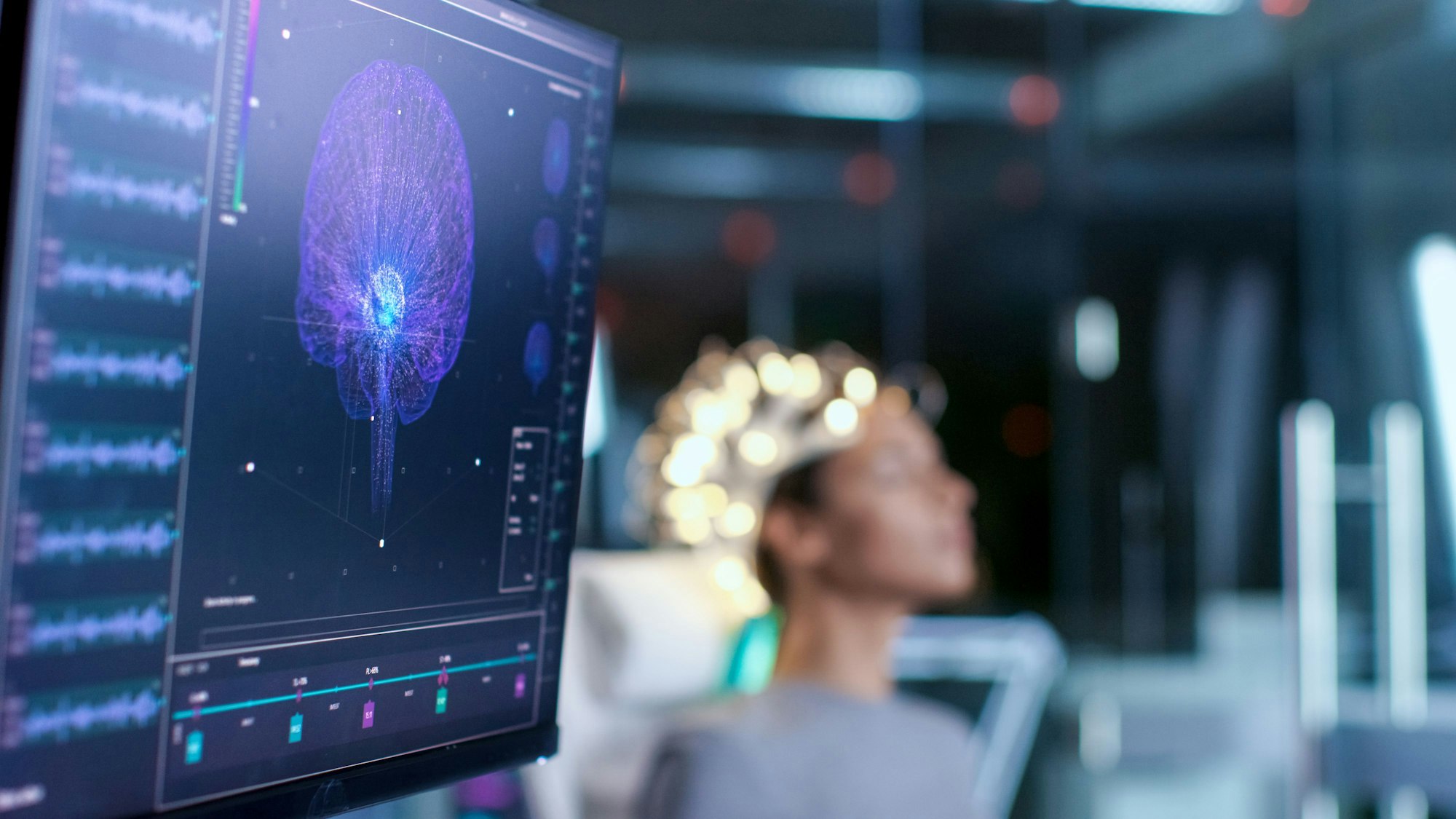 Eine Frau, die ein neurologisches Headset zum Scannen der Gehirnströme trägt, sitzt in einem Stuhl im modernen Gehirnstudienlabor/Neurologischen Forschungszentrum. Monitore zeigen EEG-Messung und Gehirnmodell.