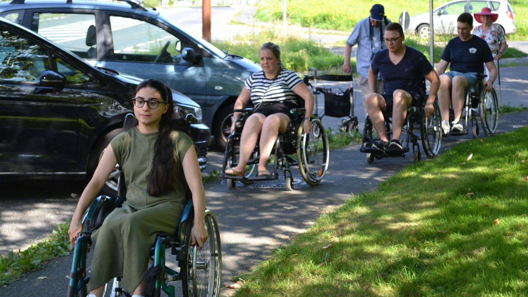 Junge Menschen in Rollstühlen passieren einen engen Gehweg.