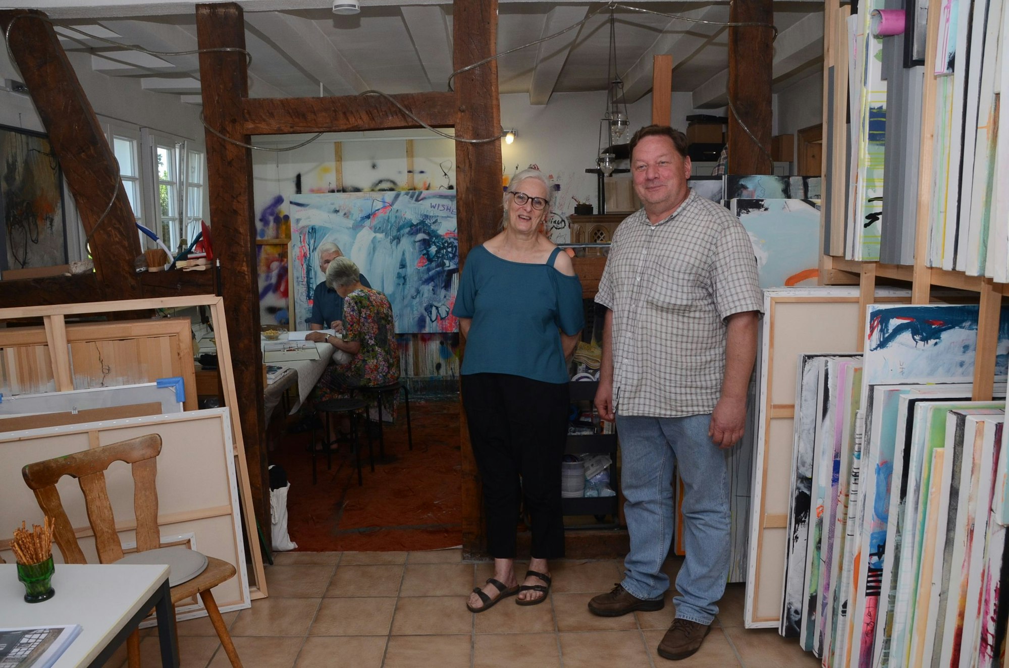 Ein Mann und eine Frau stehen in einem Fachwerkhaus. Im Hintergrund stehen Bilder auf einer Staffelei, rechts sind viele ungerahmte Bilder in einem Regal.